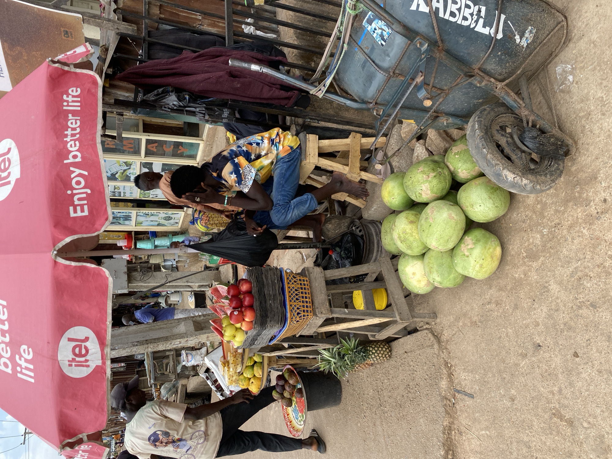 The image captures a street scene in Bukuru, Nigeria. A vendor stands beneath a pink "itel Enjoy better life" umbrella, displaying fruit including apples, mangoes, pineapples, and watermelons. A person sits on a wooden stool nearby, while another reclines on a wooden structure. A wheelbarrow laden with objects rests next to the produce.