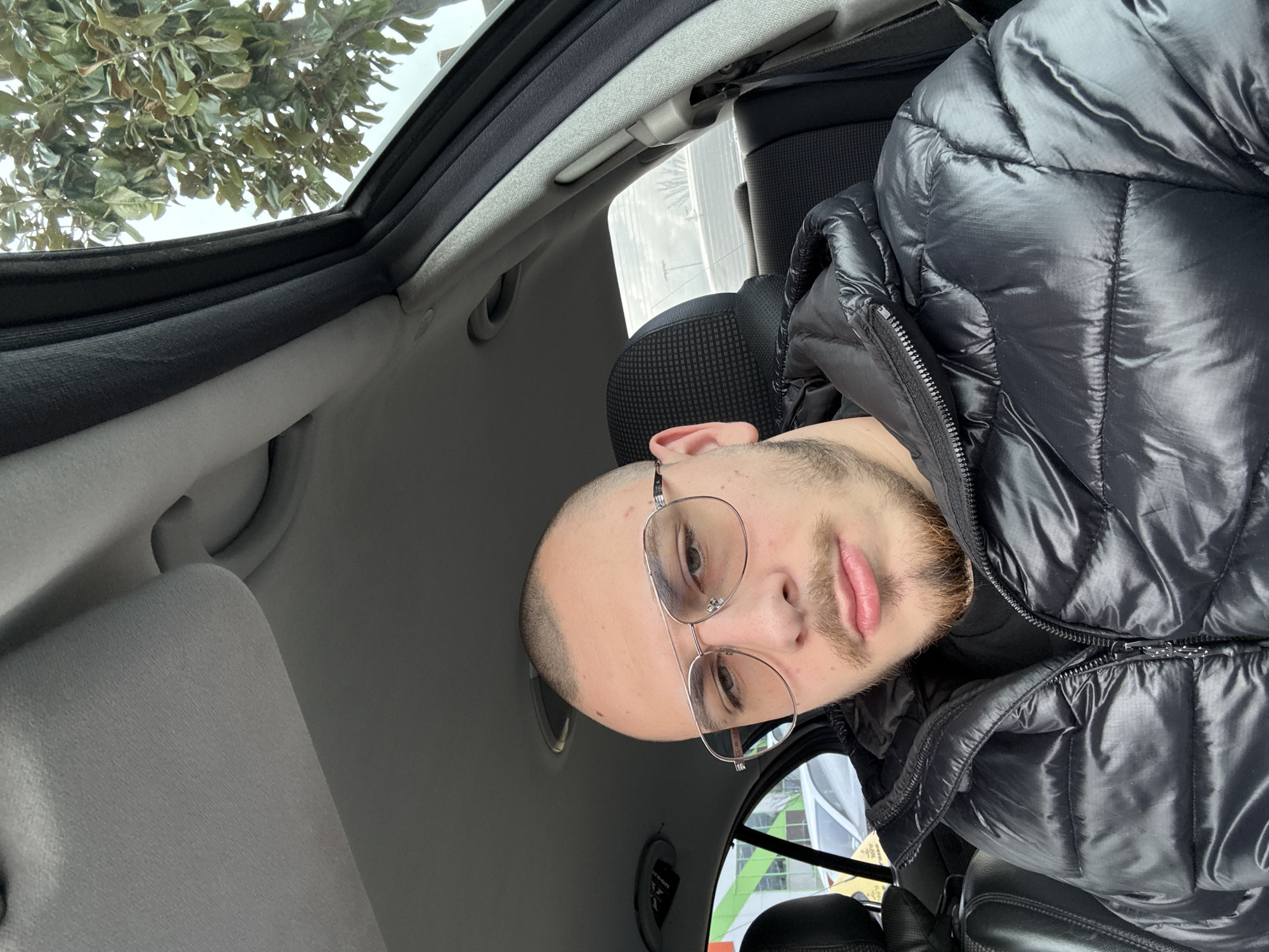 A male individual is seated in the front passenger seat of a vehicle in Stara Zagora, Bulgaria. The subject, appearing to be in his mid-twenties, has a buzzed haircut and light ginger-blonde stubble on his chin and upper lip. He wears metal-rimmed aviator-style eyeglasses with lightly tinted lenses and a black, shiny puffer jacket over a black t-shirt. His expression is neutral, with slightly pursed lips, and he is looking directly at the camera. The car's interior features a light grey headliner, sun visor, and dark grey patterned seat upholstery. Through the left-side window, a green and white striped building facade is partially visible, alongside a white vehicle. Through the right-side window, a tree with dark green leaves is seen against an overcast sky. Distant structures and a barrier are visible through the rear window.