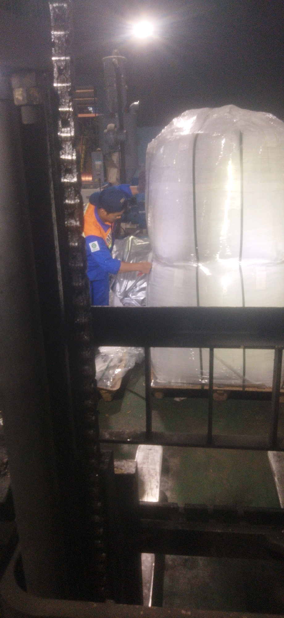 In a warehouse in Cileungsi, Indonesia, a worker wearing a blue jumpsuit and an orange vest is seen operating a forklift. The forklift's mast is visible in the foreground, partially obscuring the scene. The worker is positioned next to a large, white, cylindrical object wrapped in plastic, likely a bulk bag. The worker appears to be interacting with the wrapping on the object, possibly securing it or making an adjustment. The environment is dimly lit, with a bright overhead light source causing glare. Pallets and other industrial materials are present in the background. The scene depicts typical warehouse operations, with a focus on material handling and logistics.