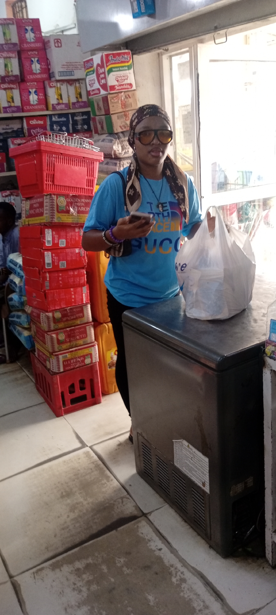The image shows a woman in what appears to be a small shop in Aviation Quarters, Nigeria. She is wearing a light blue T-shirt, dark pants, and a headscarf, and is holding a mobile phone in her hand. She is also wearing oversized sunglasses. She has a white plastic bag resting on top of a dark-colored freezer. Behind her, a stack of red plastic crates are visible, alongside boxes of pasta, juice boxes, and instant noodles ("Indomie"). The floor is tiled. A person is sitting in the background. The scene suggests a small retail establishment.