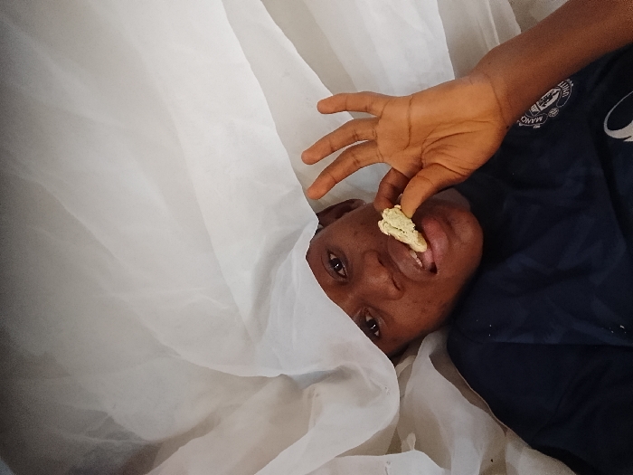 In the city of Nteje, Nigeria, an individual is depicted lying horizontally, partially obscured by a draped, translucent white fabric that covers the left side of the frame. The individual, appearing to be an adult or older child with dark skin, is wearing a dark blue shirt with a partially visible light-colored emblem on the upper left chest. Their right hand holds a small, irregularly shaped food item, light tan in color with a pale green or yellow topping, positioned between the thumb and index finger near their partially open mouth. The individual's eyes are open, directed towards the viewer. The setting appears to be indoors, likely a bed, indicated by the bedding or mosquito net.