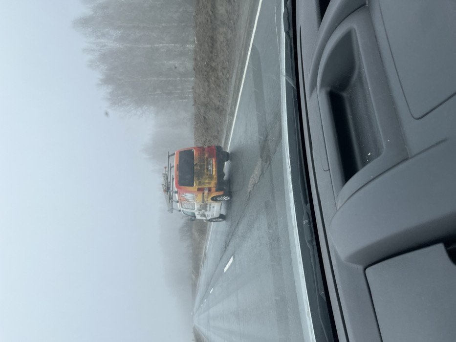 The image captures a utilitarian view from inside a vehicle traveling on a wet asphalt road in Родничок-1 Снт, Russia, during overcast or foggy conditions. The dashboard of the observing vehicle, dark in color, occupies the bottom portion of the frame. Ahead, a white utility van, prominently featuring a multi-colored orange, yellow, and brown graphic wrap on its sides and rear, proceeds in the same direction. The van is equipped with a roof rack holding multiple ladders or pipes, and its rear is soiled with dirt or mud. The two-lane road exhibits visible cracks and potholes, with white dashed and solid lane markings. To the right, a raised dirt embankment parallels the road, beyond which a line of bare, deciduous trees extends into the obscured background due to fog or atmospheric haze. Similar indistinct tree lines are visible further in the distance. No individuals are discernible.
