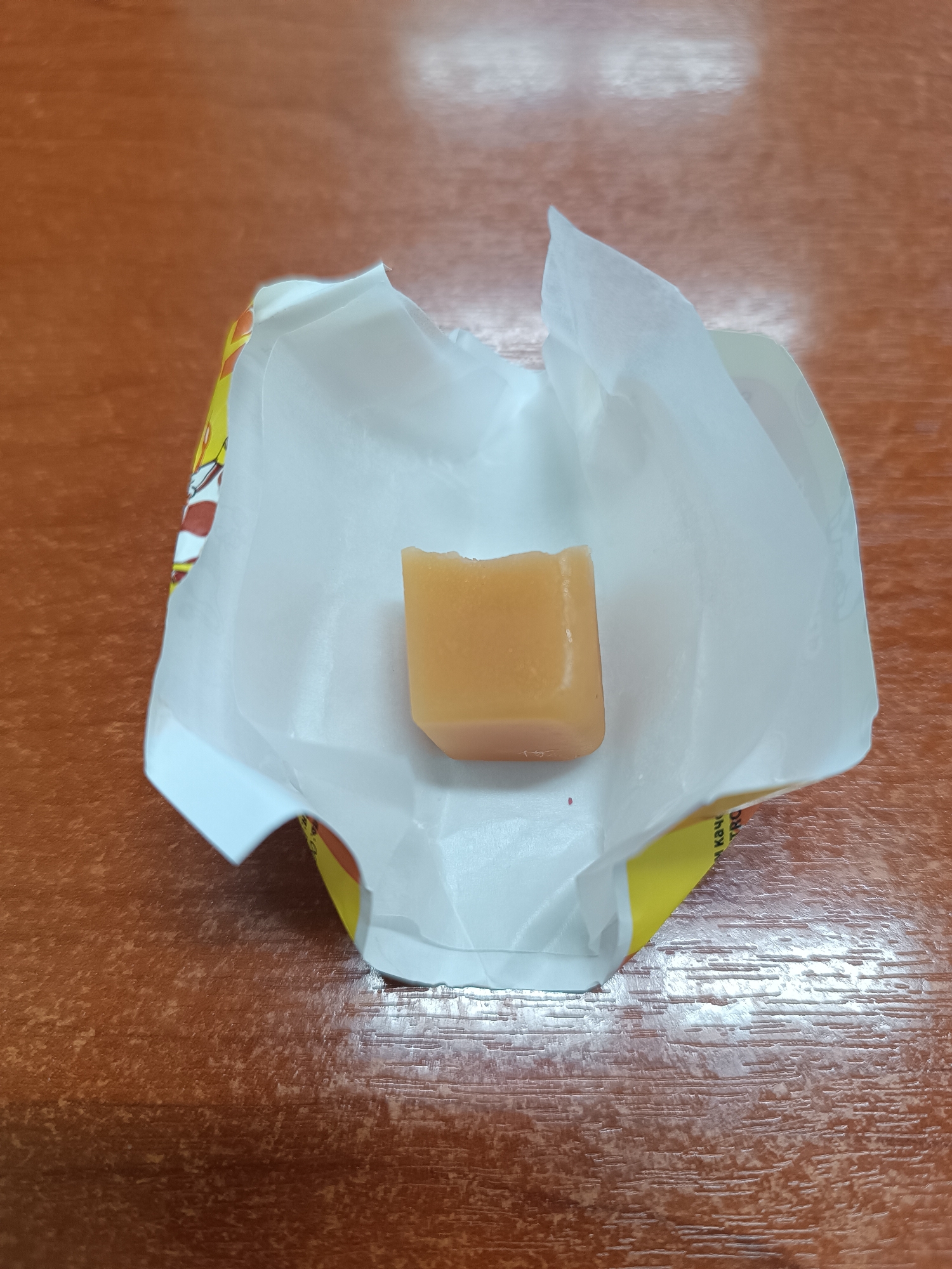 The image shows a partially unwrapped caramel candy on a table. The candy is light brown and appears to have a bite taken out of it. It sits inside its opened wrapper, which consists of a white inner layer and a yellow outer layer with some text in a foreign language and a cartoon image. The table has a wood grain pattern. The image was taken in Chaikovsky, Russia.