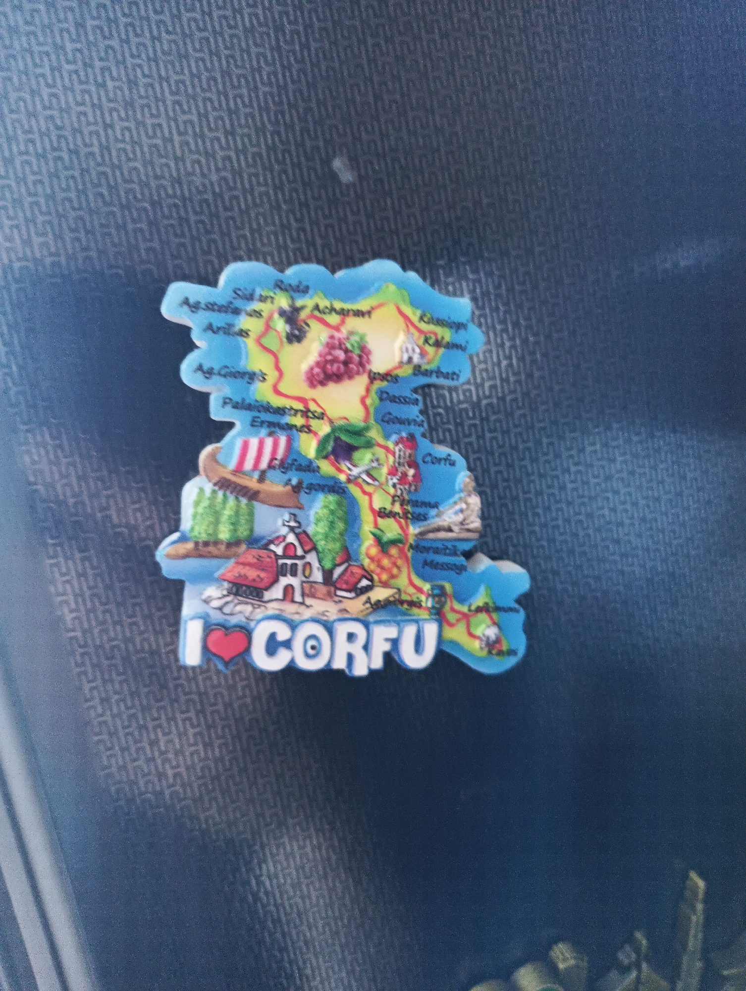 This is a close-up, high-angle shot of a novelty magnet shaped like the island of Corfu. The magnet is a colorful, stylized map of the island with various towns and landmarks indicated by text and small illustrations. At the bottom of the magnet, the phrase "I ❤️ CORFU" is written in large white letters with a red heart in place of the word "love." The magnet is lying on a dark, textured surface, possibly a fabric or patterned material. The lighting appears to be overhead, creating some shadows and reflections on the surface. There are no people or animals present in the image, and the focus is solely on the magnet. The image does not depict the city of Karnobat, Bulgaria.