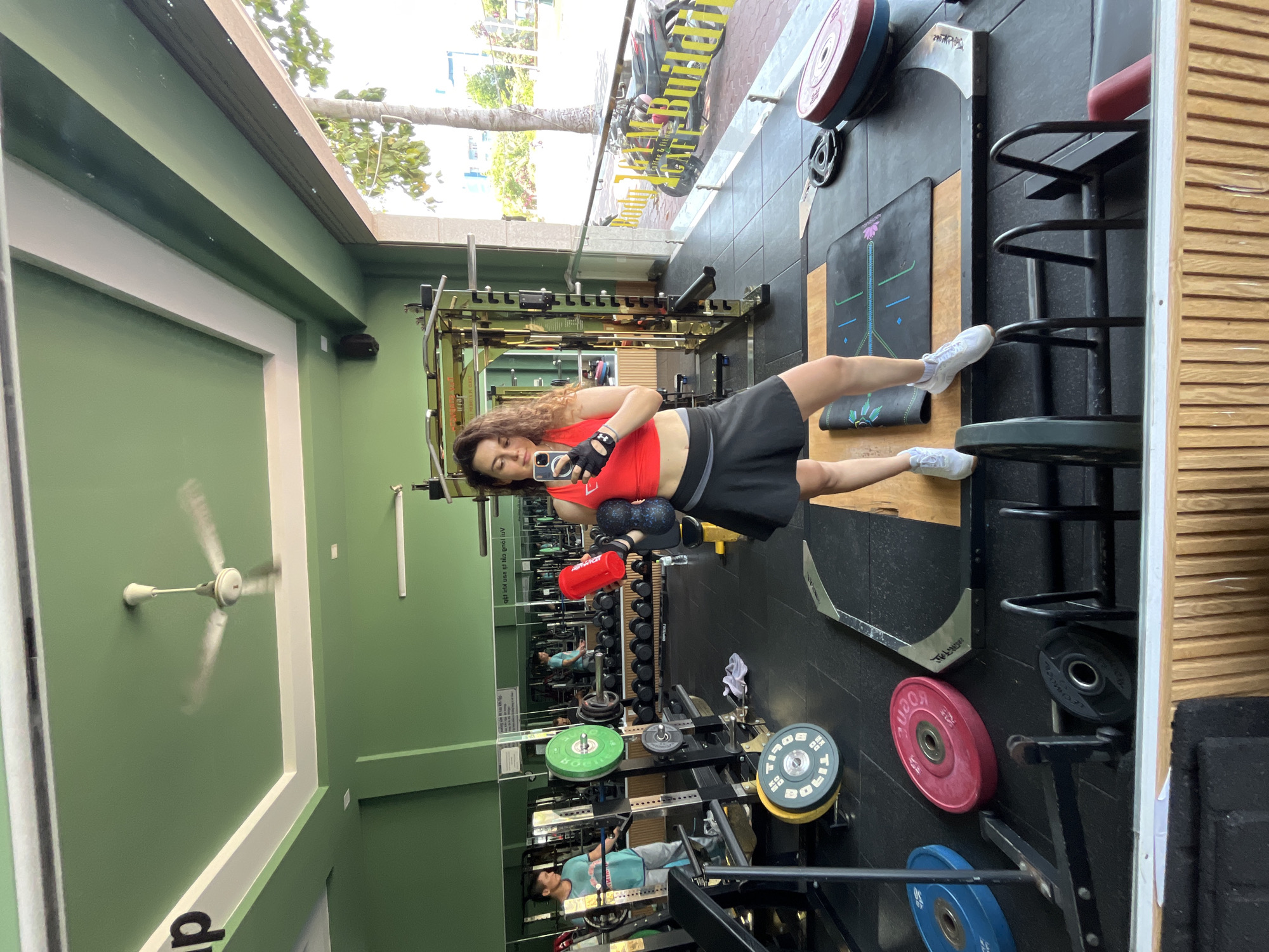 A woman is taking a mirror selfie in a gym in Phan Thiết, Vietnam. She is wearing a red crop top, black skirt, and white sneakers, and is holding a red bottle and a black foam roller. The gym has green walls, a white ceiling with a ceiling fan, and various gym equipment, including weight racks, barbells, and weight plates. In the reflection, another person can be seen in the background using a weight machine. The gym has large windows that look out onto a street with trees and buildings. The overall scene depicts a fitness-oriented environment.