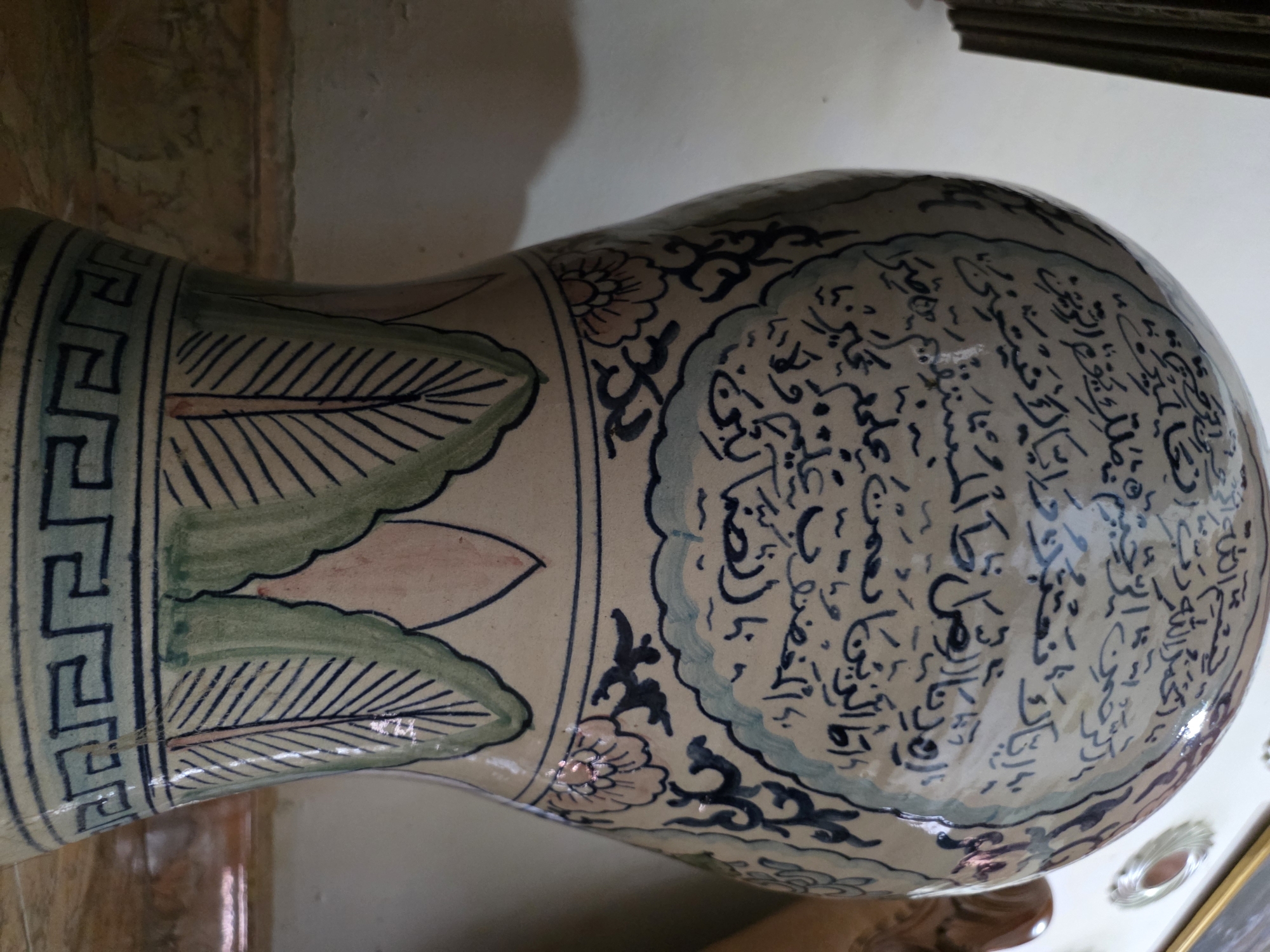 The image shows a close-up of a decorative ceramic vase with Arabic calligraphy. The vase is primarily off-white with blue and green painted details. The calligraphy, in dark blue, covers a significant portion of the upper section of the vase, arranged within a cloud-like border. Below this, there are floral motifs and stylized green leaf designs, followed by a geometric pattern along the base. The vase appears to be an interior decorative object. No specific location in Bireuenmeunasahblang, Indonesia can be identified from the provided image.
