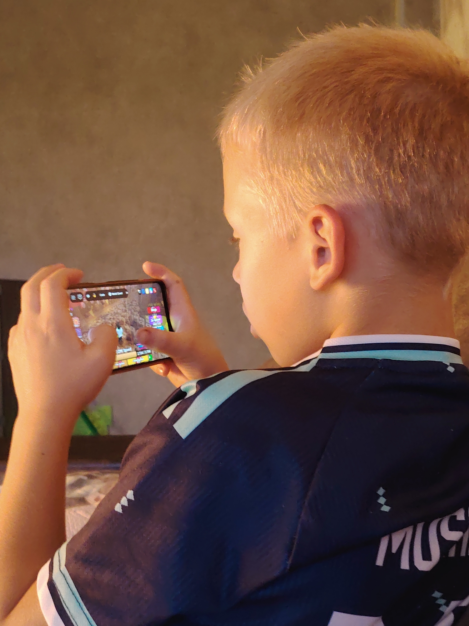 A blonde boy is playing a video game on a smartphone. He is holding the phone horizontally with both hands, his thumbs touching the screen. The phone screen shows a colorful game with characters and a map. The boy is wearing a dark blue t-shirt with light blue and white stripes on the collar and sleeves. The background is a plain, textured wall. The lighting suggests the image was taken indoors with natural light coming from the side. There is no discernible information about the specific location in the city of Chaikovsky, Russia.