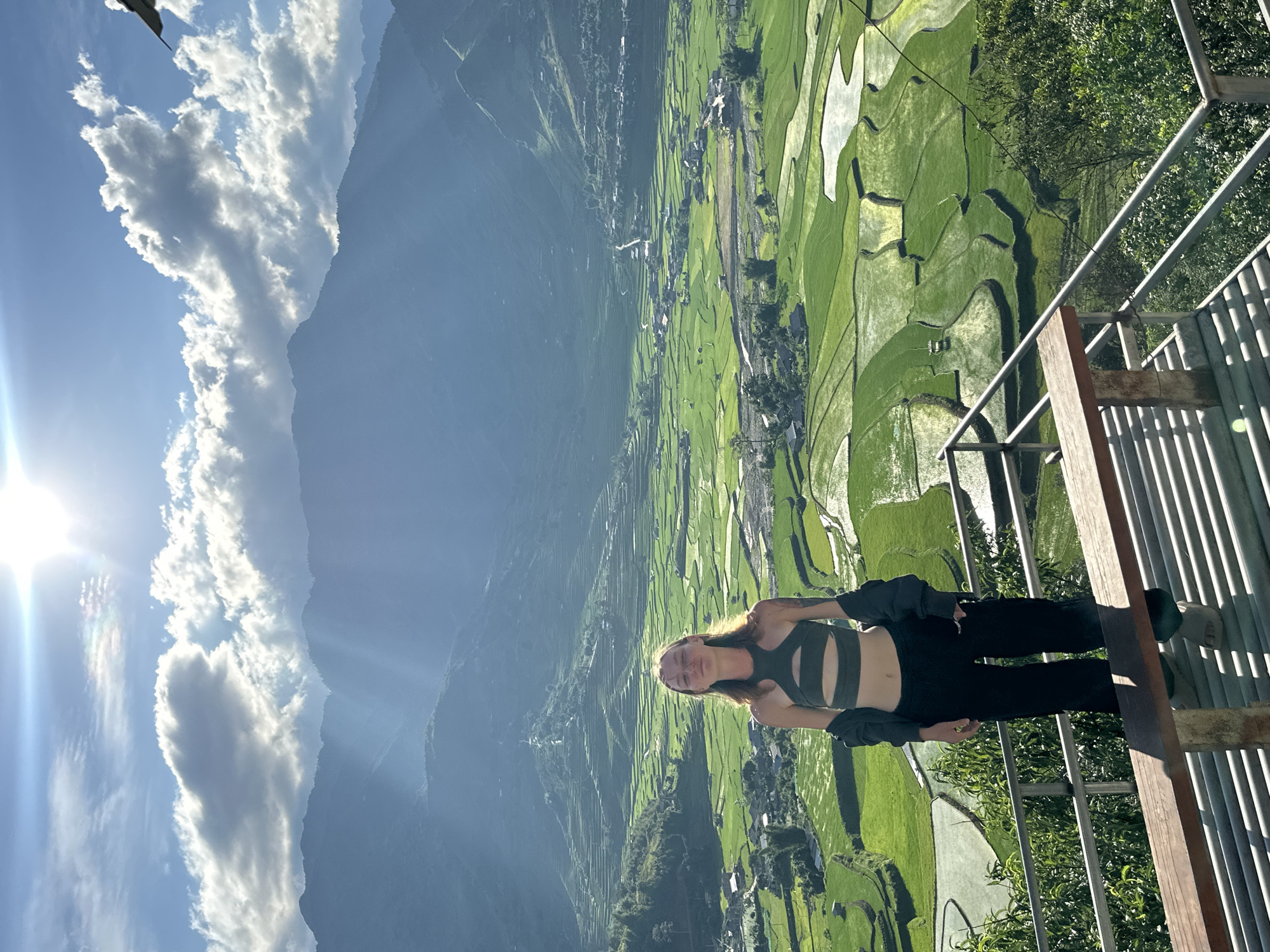A young adult female stands on a metal grate platform with a wooden bench, facing the camera with a slight smile and closed eyes, against a panoramic backdrop of terraced rice fields in Tú Lệ, Vietnam. She wears a dark olive-green cut-out crop top, dark trousers, and a dark jacket draped over her shoulders, with light-colored sandals. A tattoo is visible on her left arm. The vibrant green rice paddies, some reflecting water, spread across a valley floor and ascend the surrounding mountainsides, interspersed with clusters of houses and a winding road. Distant mountains rise under a bright blue sky with scattered white cumulus clouds, and a strong sun in the upper right casts rays and causes lens flare, backlighting the scene and creating atmospheric haze over the distant peaks. Metal railings define the edge of the foreground platform.