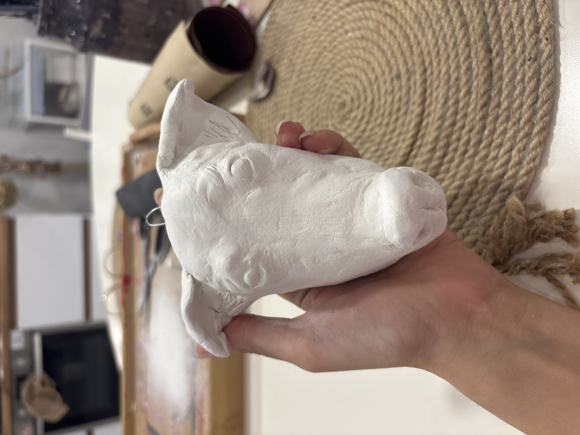 A human hand holds a roughly sculpted, white clay animal head. The animal is not clearly defined but appears to be in the style of a cartoon pig or similar mammal. The hand is light-skinned. The background is out of focus and cluttered with objects including a roll of brown paper or cardboard, a coiled rope mat, and a blurry view of items suggesting an art studio. The location is not apparent from the visual details provided. There is no evidence of it being in Krasnoyarsk, Russia.