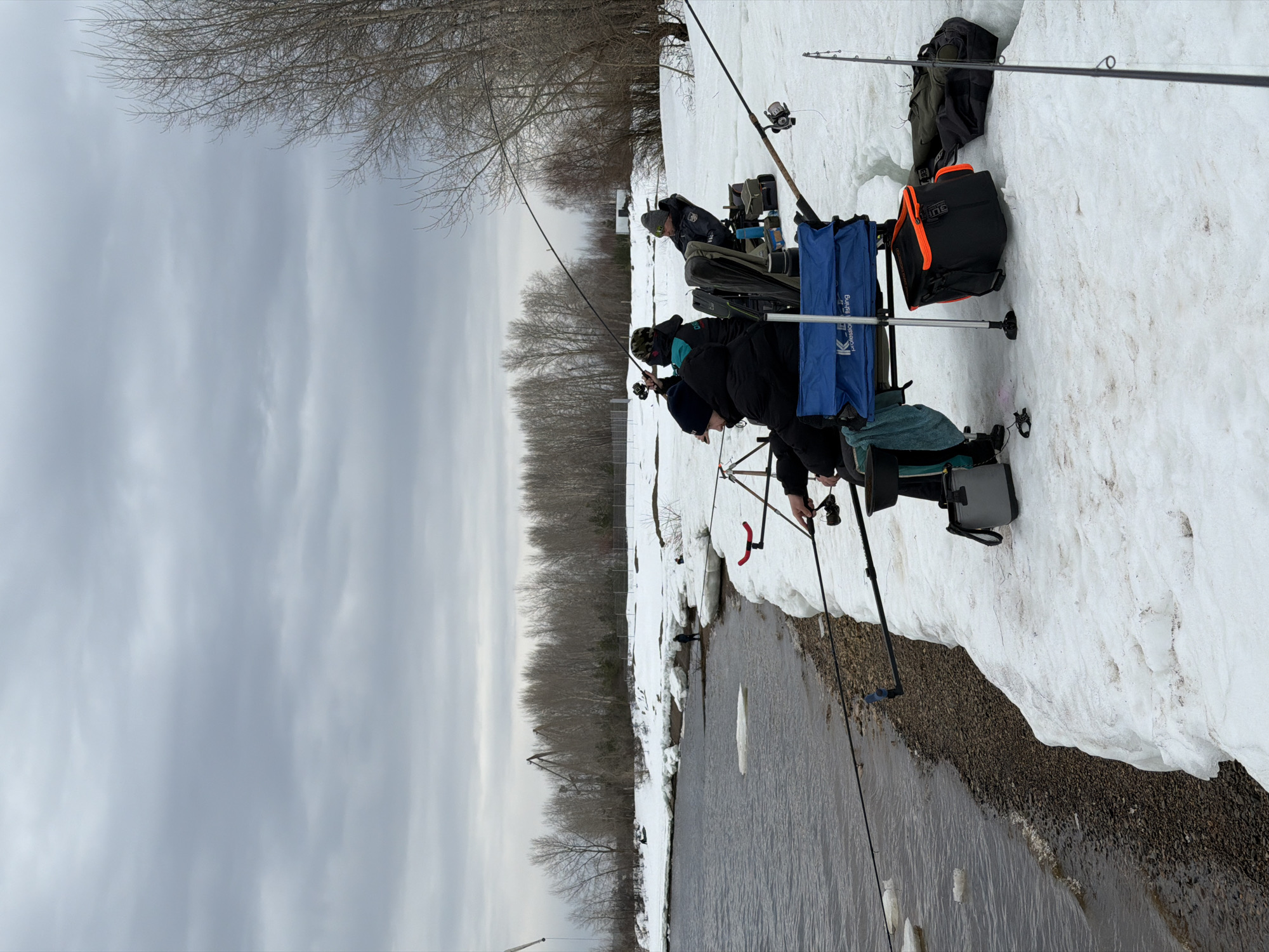 The image depicts three individuals fishing along the icy edge of a riverbank. The location is suggested to be Noviy, Russia. Patches of snow cover the ground around them, extending to the river's edge. Each person has a fishing rod, with some rods angled out over the water. Fishing equipment and bags are positioned near them on the snow-covered ground. The river flows through the scene, and bare trees line the opposite bank, reaching towards a cloudy sky.
