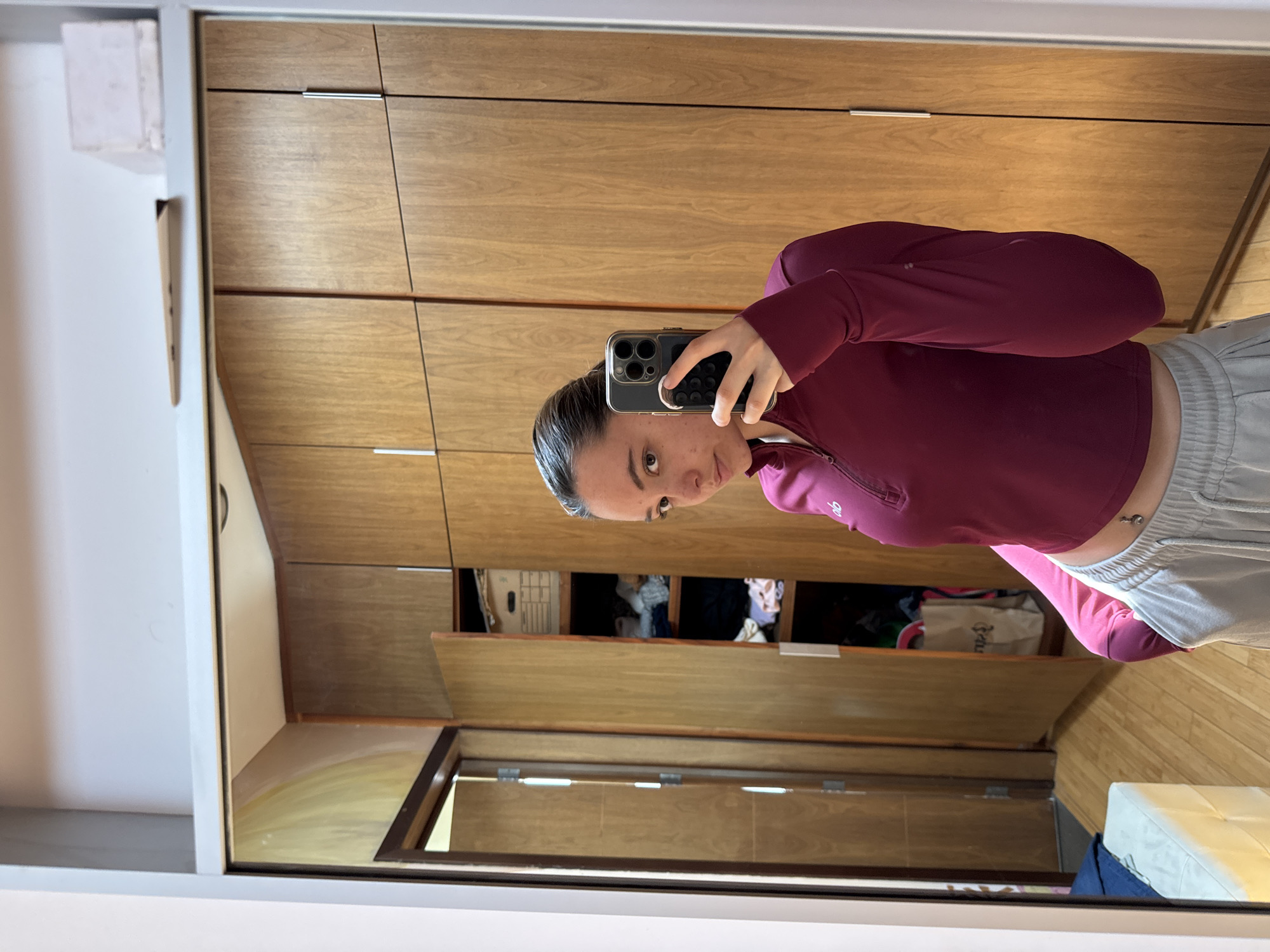 A person is captured in a mirror selfie, holding a smartphone with a distinctive multi-lens camera system and a textured grip on the back. The individual is positioned centrally in the frame, with their head slightly tilted and a direct gaze towards the camera. Their dark hair is pulled back, and they appear to have minimal makeup.
They are wearing a long-sleeved, cropped athletic top in a dark magenta or burgundy color, featuring a quarter-zip neckline. The brand "alo" is visible on the left chest area of the top. Paired with this, they wear light grey sweatpants or athletic shorts with a drawstring waist. A belly button piercing is visible.
The setting is an indoor space, reflected in a large mirror. Behind the person, a series of light wooden cabinets or wardrobes with horizontal paneling and slender silver handles are visible. Some of these cabinet doors are open, revealing stored items such as folded clothes and a light-colored bag with text that appears to start with "Ru". In the lower right corner of the reflection, a white, cushioned object, possibly a stool or ottoman, is partially visible. The overall lighting suggests daytime.
The image is taken in San Bernardino Tlaxcalancingo, Mexico.