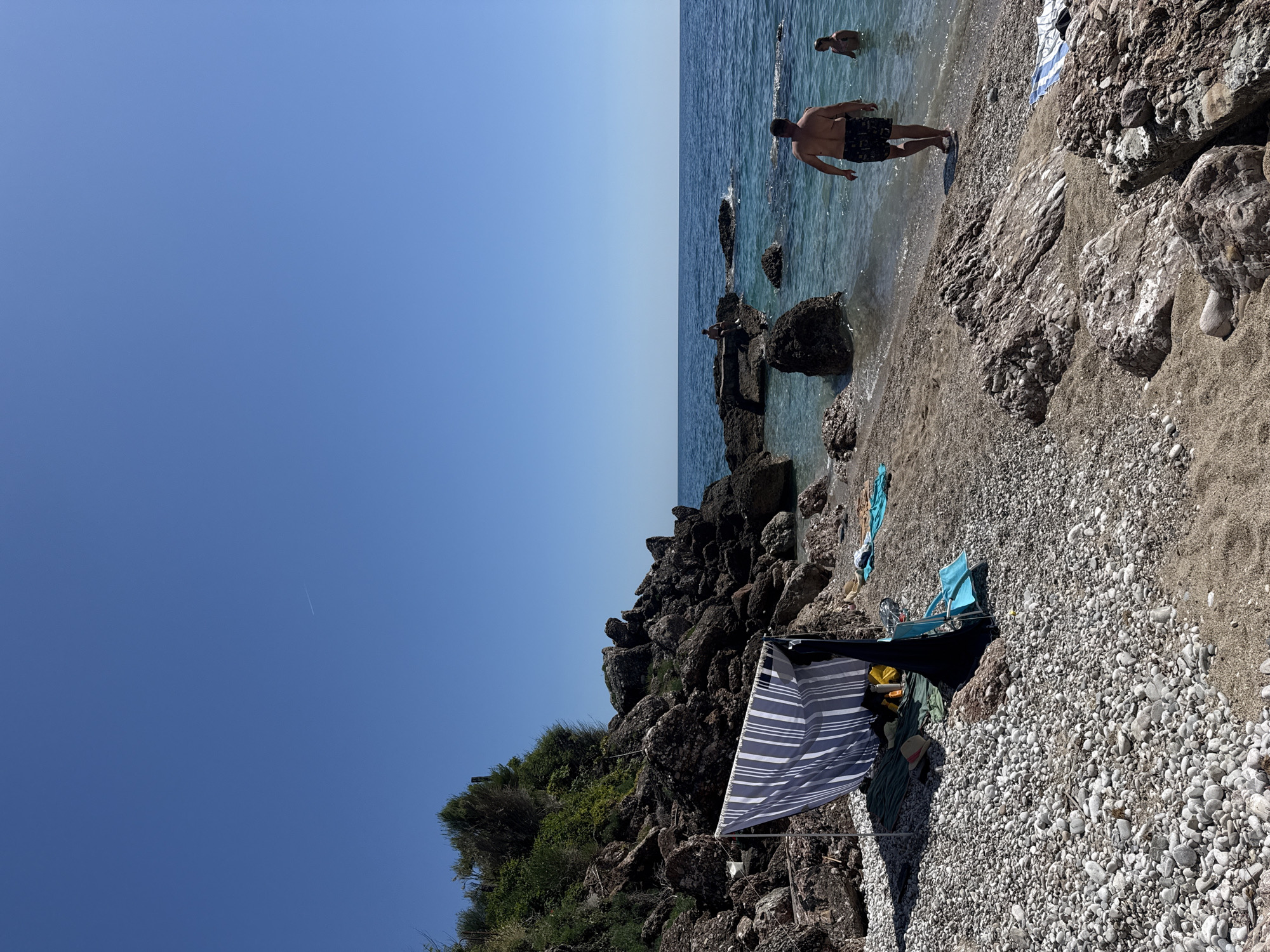 This image depicts a sunny day on a rocky and pebbly beach in Drobnići, Montenegro. The scene is dominated by a clear blue sky, featuring a faint white contrail in the upper mid-right, and a calm, turquoise-blue sea.
On the left side of the beach, a blue and white striped windbreak is erected, providing shade. Beneath it, a green mat is laid out, holding a yellow bag, dark clothing, and a black bag. A light blue folding beach chair is positioned beside the windbreak. The ground consists of numerous grey and white pebbles that transition to coarser sand closer to the water.
Towards the center-right, a man with his back to the viewer, wearing dark patterned swim shorts, walks into the clear water. To his right, another individual is partially submerged in the sea. Further out, a person is seated on a large, dark rock formation that rises from the water.
The left periphery of the image features a rugged hillside composed of dark rocks and boulders, densely covered with green vegetation and shrubs. Various large rocks are scattered along the shoreline and partially submerged in the shallow water. The horizon line, separating the sea from the sky, is visible across the upper portion of the image.