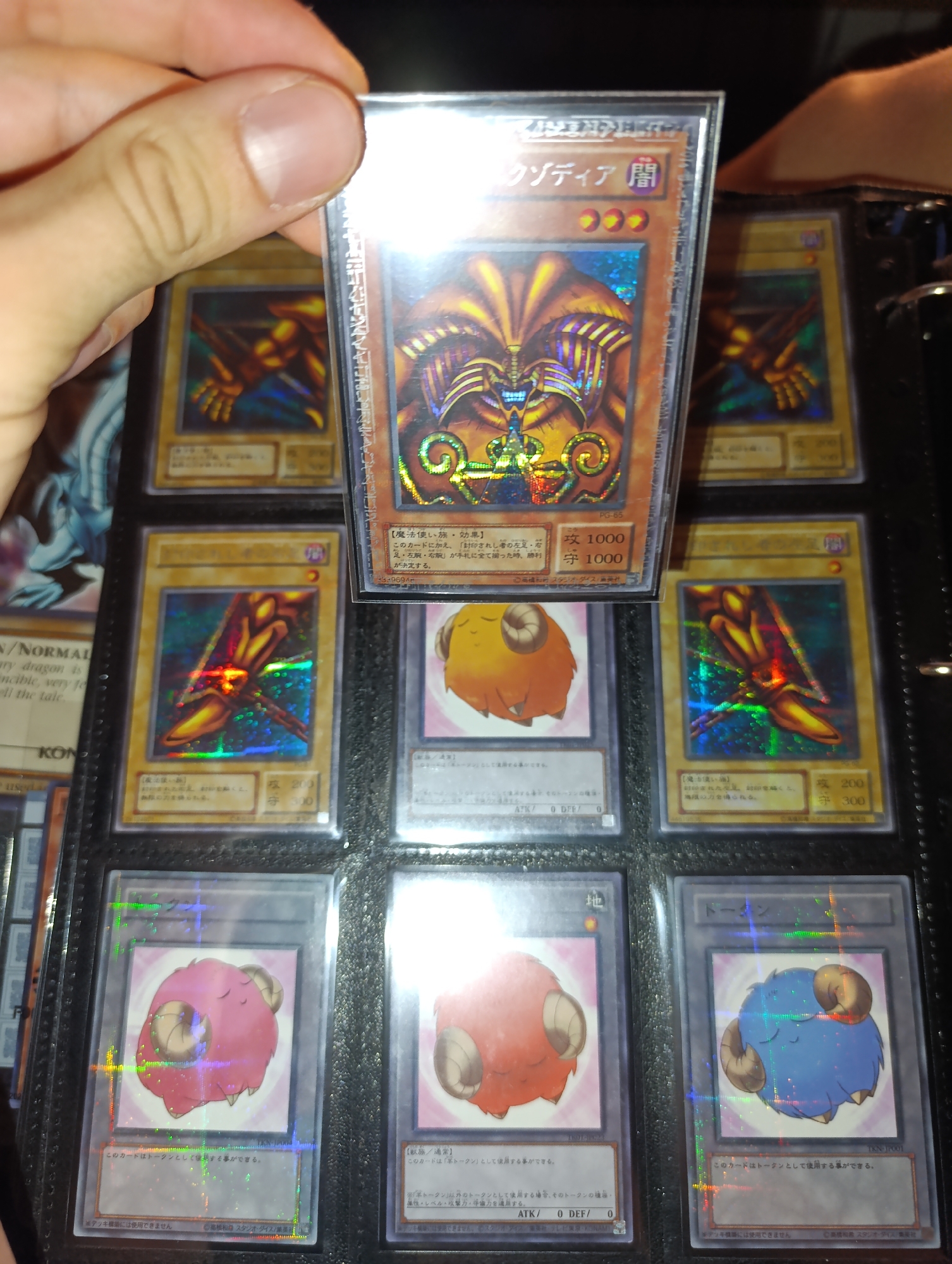 The image shows a collection of Yu-Gi-Oh! trading cards, displayed in a binder page. In the upper row, there are two Exodia cards and a partially visible third one. In the row below, there are two Exodia pieces and a Sheep Token card. In the lower row, there are three Sheep Token cards of different colors (pink, red and blue). A hand is holding a holographic Exodia Necross card in front of the binder. The cards are sleeved and placed in the binder pockets. Some cards show holographic patterns. The focus is on the cards, their arrangement, and the hand holding one of them.