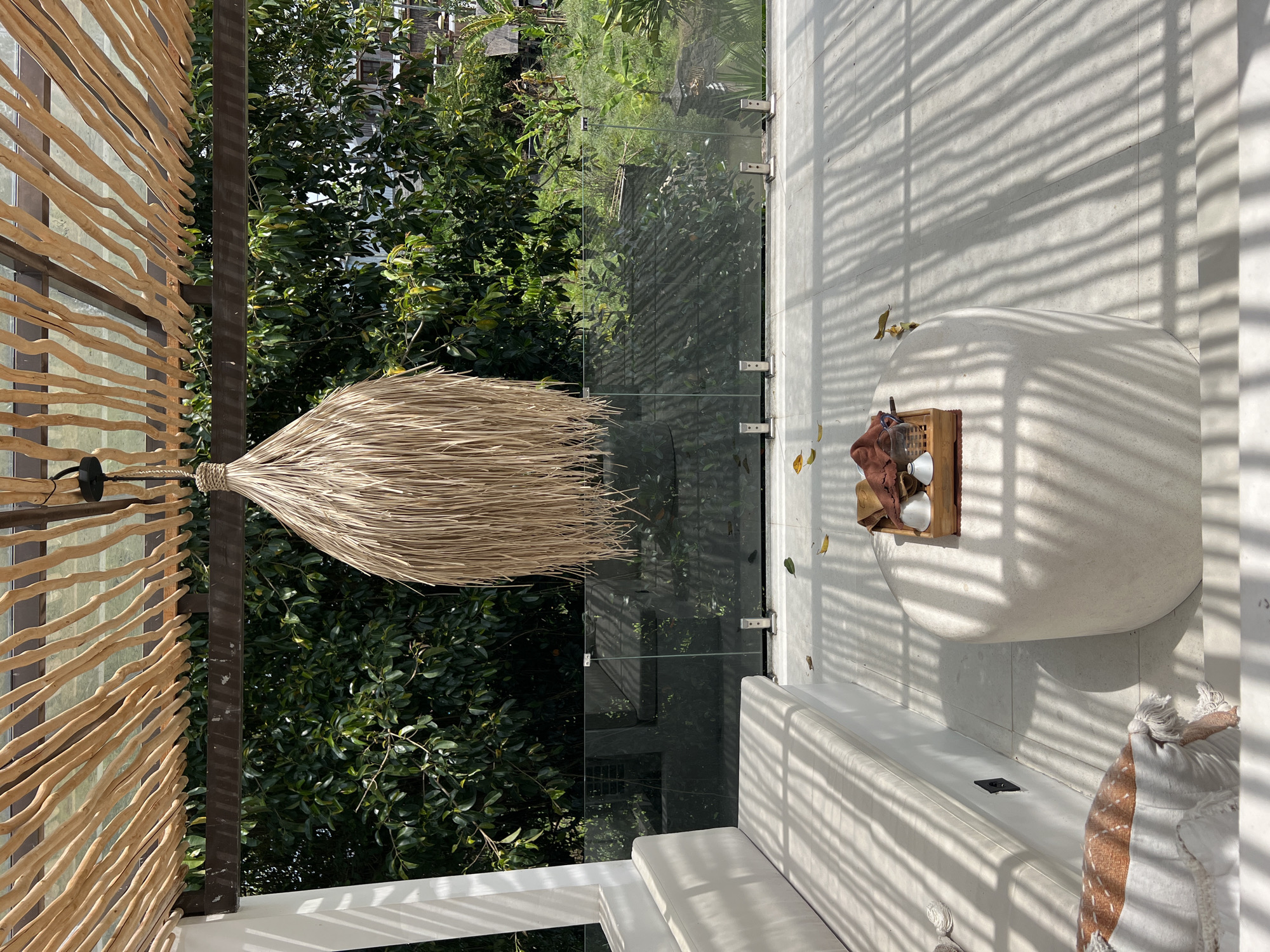 The image depicts a terrace in Buduk, Indonesia. A decorative lamp made of straw hangs from a wooden slat wall, which is constructed using dark wood. A glass railing separates the terrace from a lush green garden. Inside the terrace, the flooring is light-colored tile covered in shadows from an unseen structure. A low white sofa sits against the glass railing, and a round white table rests on the tile flooring. A wooden tray on the table holds cups and a red cloth. Next to the sofa is a cushion.