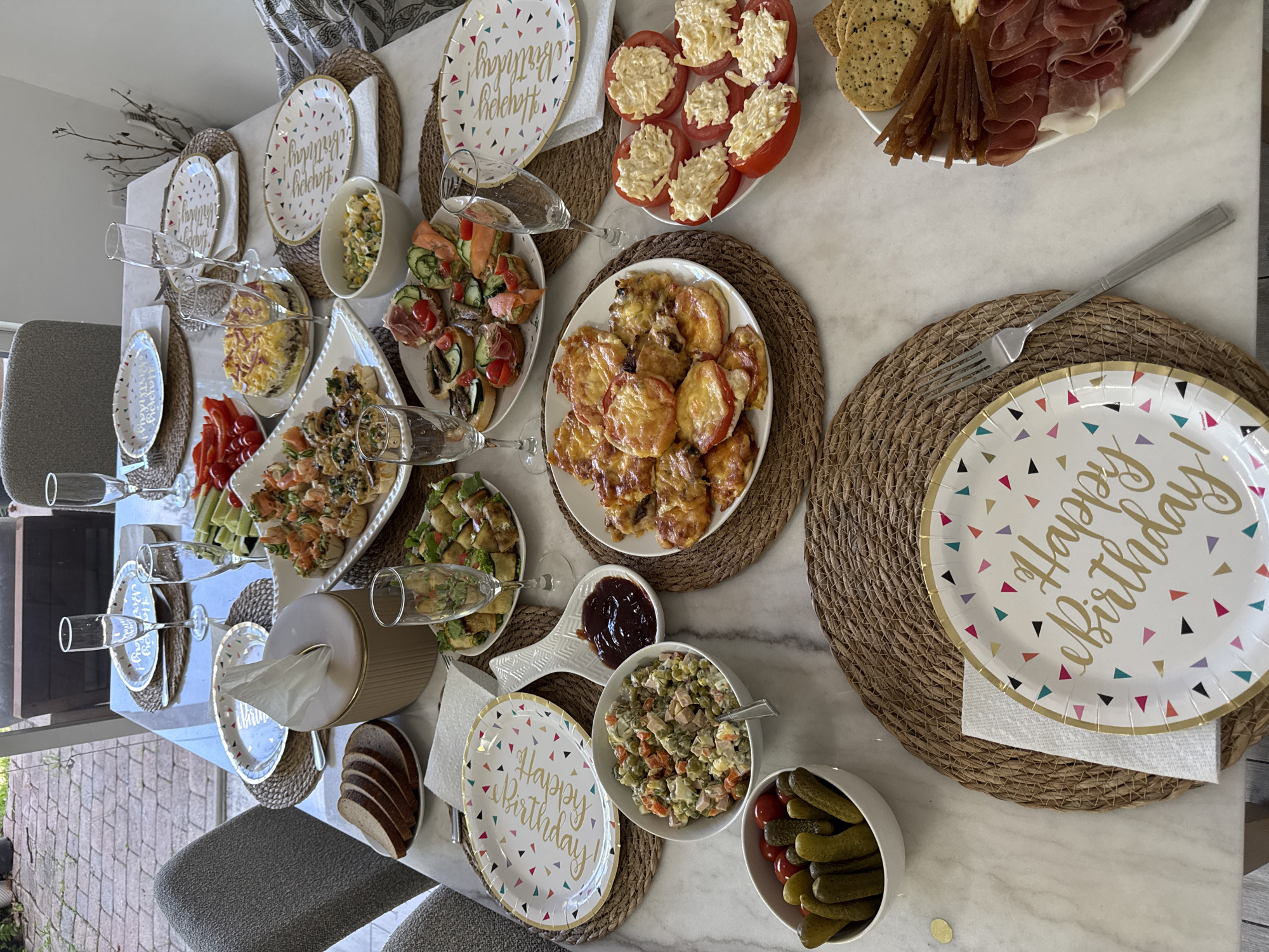 A long dining table, set for a celebratory meal, is presented from a top-down perspective in an indoor setting with a view of a patio through a large window. The table is covered with a marble-patterned tablecloth and adorned with various dishes, plates, and glassware, suggesting a birthday party or special occasion.
Several plates with a "Happy Birthday" design, featuring colorful confetti, are placed on woven placemats. To the left of the plates, stacks of sliced brown bread are visible, along with a tissue box. Various appetizers are spread across the table, including shrimp appetizers in small pastry shells, mini sandwiches with salmon and other toppings, and a salad composed of peas, carrots, potatoes, and ham. There is also a dish of sliced tomatoes topped with a creamy filling and a plate of cured meats and crackers. A small bowl of pickles and cherry tomatoes sits on a placemat.
Champagne flutes are placed at intervals around the table, some empty and some appearing to have contained liquid. A decorative spoon holds a dark red condiment, likely jam or sauce. The lighting suggests it is daytime, and the overall atmosphere is convivial and festive. The scene is set within a home in Dania Beach, Florida, United States of America, evidenced by the specific geographical location provided for this task. No people are visible in the image.