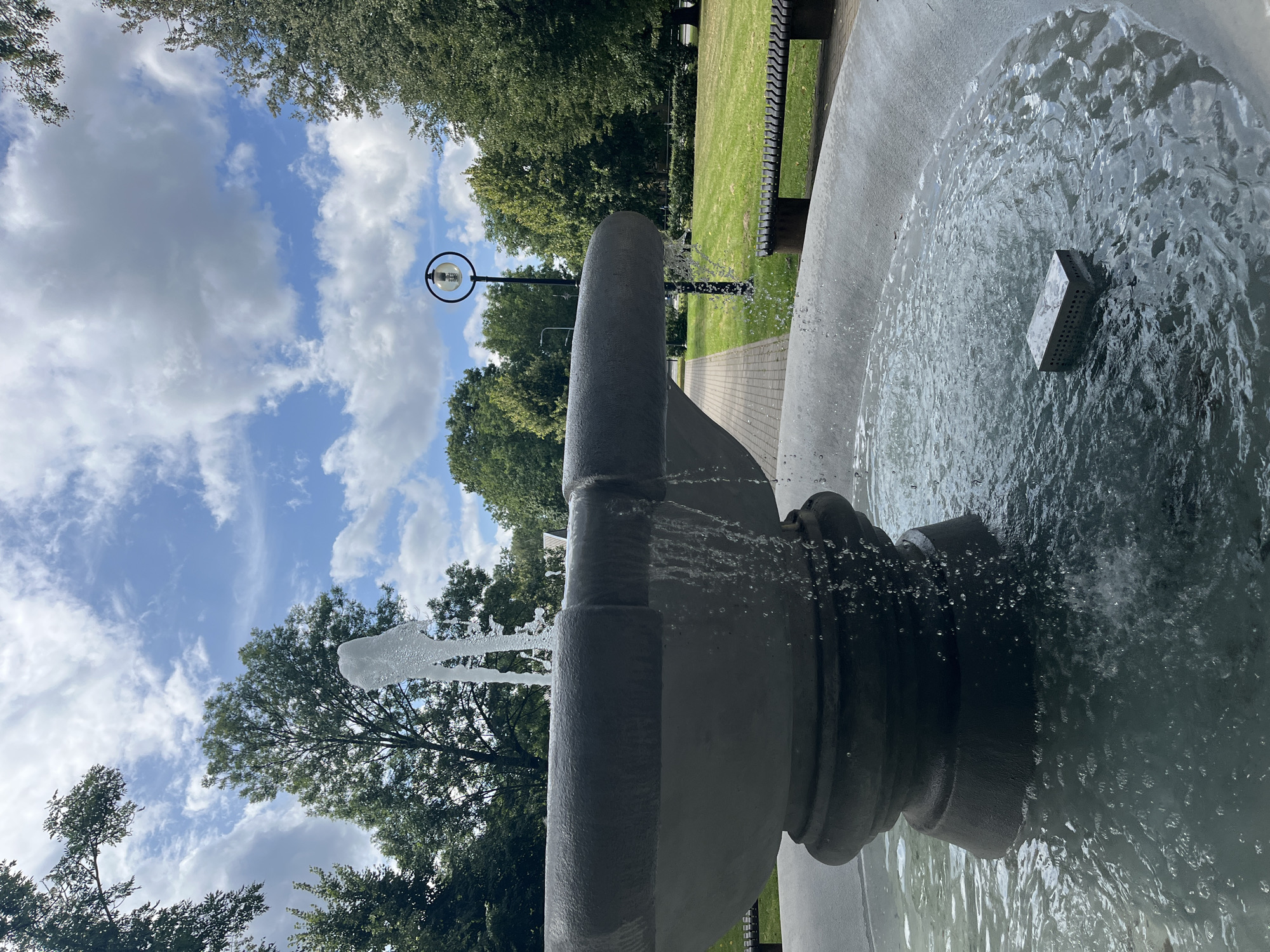 The image shows a stone fountain in an outdoor park setting. The fountain has multiple tiers and water is actively flowing from the top, creating a spray and cascading down into the basin. In the basin, there is a rectangular object with holes in it floating on the surface of the water.
Behind the fountain, a paved walkway leads into the distance, bordered by a well-maintained grassy area. Park benches are visible along the edge of the lawn. A lamppost with a circular design is situated further down the walkway. Lush green trees surround the park, and the sky above is a bright blue with scattered white clouds.
The specific location is identified as the city of Kohtla-Järve linn, Estonia, suggesting this is a public park within that city. The scene appears peaceful and well-kept, indicating a public space designed for recreation or relaxation. There are no people visible in the image.