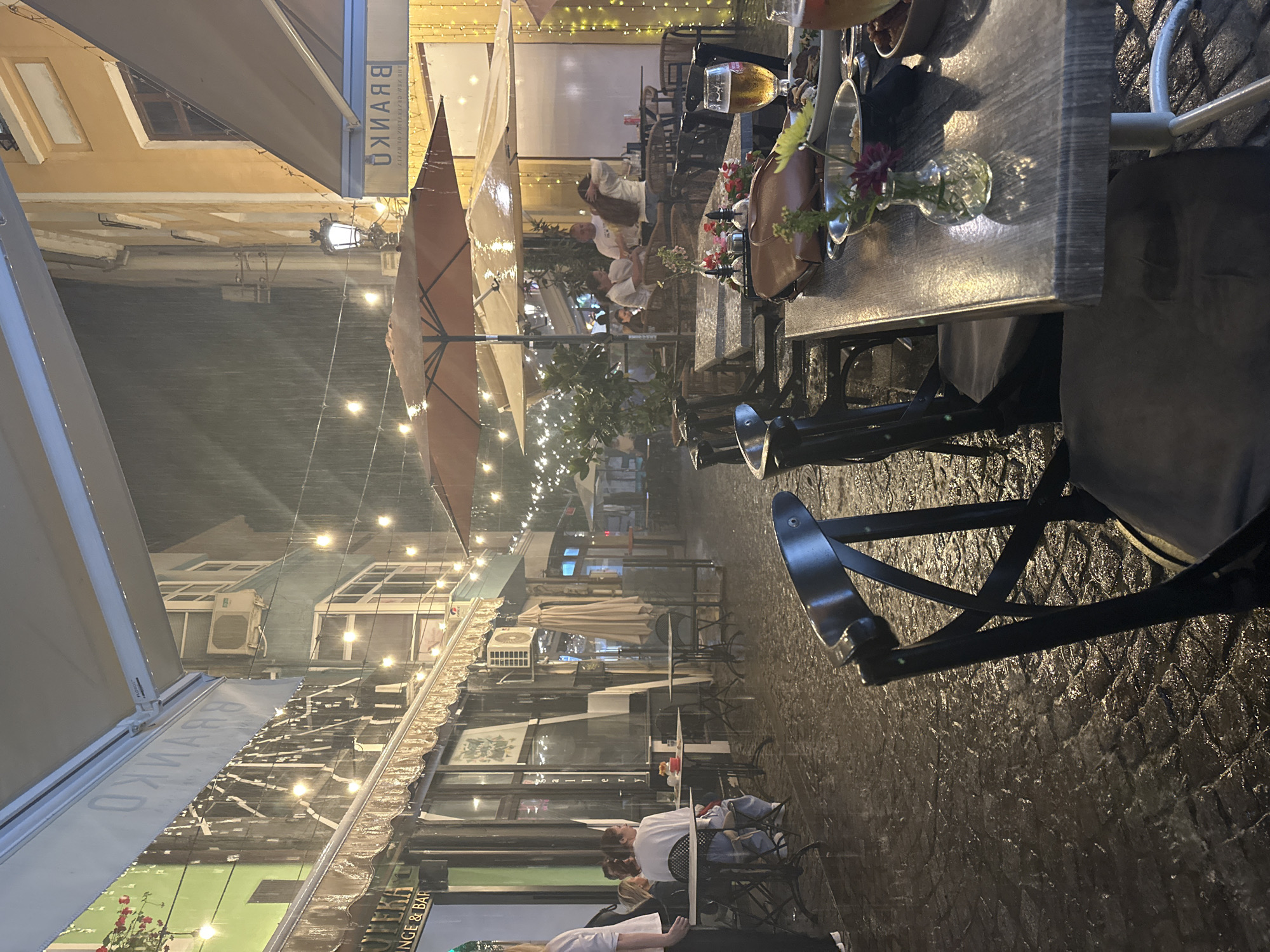This detailed image captures an outdoor dining scene on a cobblestone street in the Kapana district of Plovdiv, Bulgaria, specifically in front of "Potteka Lounge & Bar" and "Branko Lounge & Bar," during a nocturnal rain shower.
In the immediate foreground on the right, the back of a black wooden chair with a padded seat is visible. To its left is a wooden table with a light-colored surface, upon which a small, clear glass vase holding a single dark red flower is placed. Other items on this table include a brown leather purse, a small metal bowl containing food (possibly a dip or spread), another bowl with dark contents (possibly olives), and a large glass of golden beer with foam. Cutlery and another partially visible glass are also present. The cobblestone ground beneath is visibly wet and reflective from the rainfall.
The midground features numerous outdoor tables, chairs, and several large, beige patio umbrellas, some partially closed or dripping with water. Rain streaks are evident throughout the scene, illuminated by numerous warm string lights strung overhead between buildings. On the right side of the midground, three individuals (two women and one man) are seated around a table under an umbrella. One woman in a white top is seen adjusting her long hair. Further left, another woman in a white top and blue jeans is seated at a table facing away from the camera, with another person partially visible next to her. A person in a white shirt and dark pants stands partially cropped on the far left.
The background on the left reveals a building with a green facade. An awning prominently displays "POTTEKA LOUNGE & BAR" and, further back, the word "BRANKO" in white, mirrored lettering. Hanging planters with red flowers are visible. An external air conditioning unit is mounted on the building. On the right background, a building with an ochre-yellow facade is seen, featuring windows with dark frames. An awning on this side also displays "БРАНКО" (Branko in Cyrillic), mirrored. A traditional-style street lamp with an ornate base and a glowing bulb is positioned between the buildings. The wet cobblestone street extends into the distance, reflecting the artificial lights.