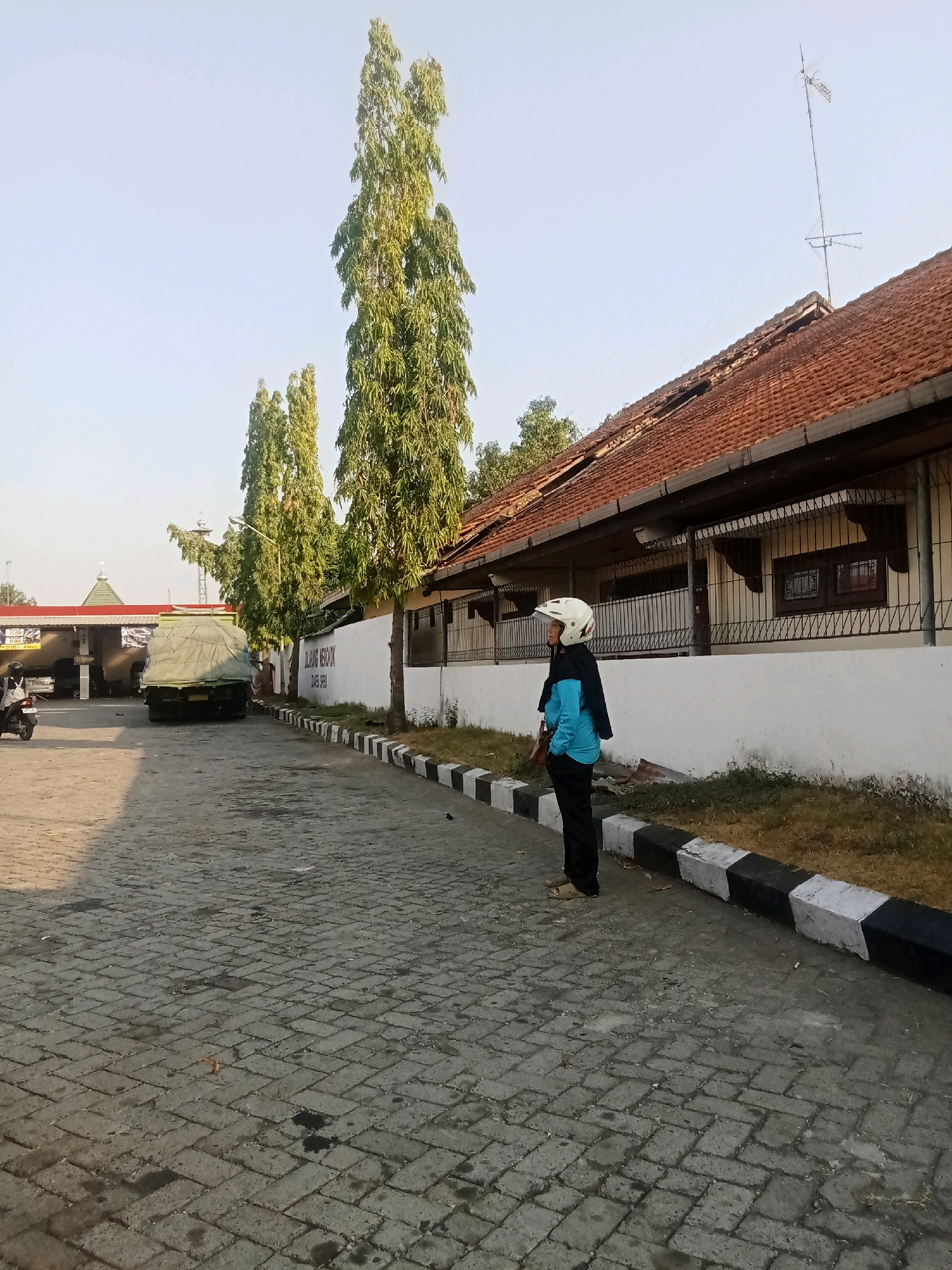 The image depicts a street scene in the city of Jelakombo, Indonesia, on a clear day. The foreground features a paved road with interlocking brick pavers, showing some signs of wear and moisture. A person wearing a blue shirt, dark pants, and a white helmet is standing on the sidewalk, facing to the left. The sidewalk is bordered by a curb painted with alternating black and white blocks.
To the right, a building with a terracotta tile roof and white walls is visible. The building has windows with decorative iron grilles. An antenna is mounted on the roof. Behind the building, tall, slender green trees rise towards the sky.
In the mid-ground, a large truck covered with a green tarp is parked on the road. To the left of the truck, there appears to be a covered structure with signage and some vehicles parked underneath, possibly a gas station or service area. Further in the background, more trees and structures are visible. A motorcycle is parked on the left side of the frame, with a person on it. The overall environment suggests an outdoor, possibly commercial or roadside area, with a low angle of the sun indicating either early morning or late afternoon.