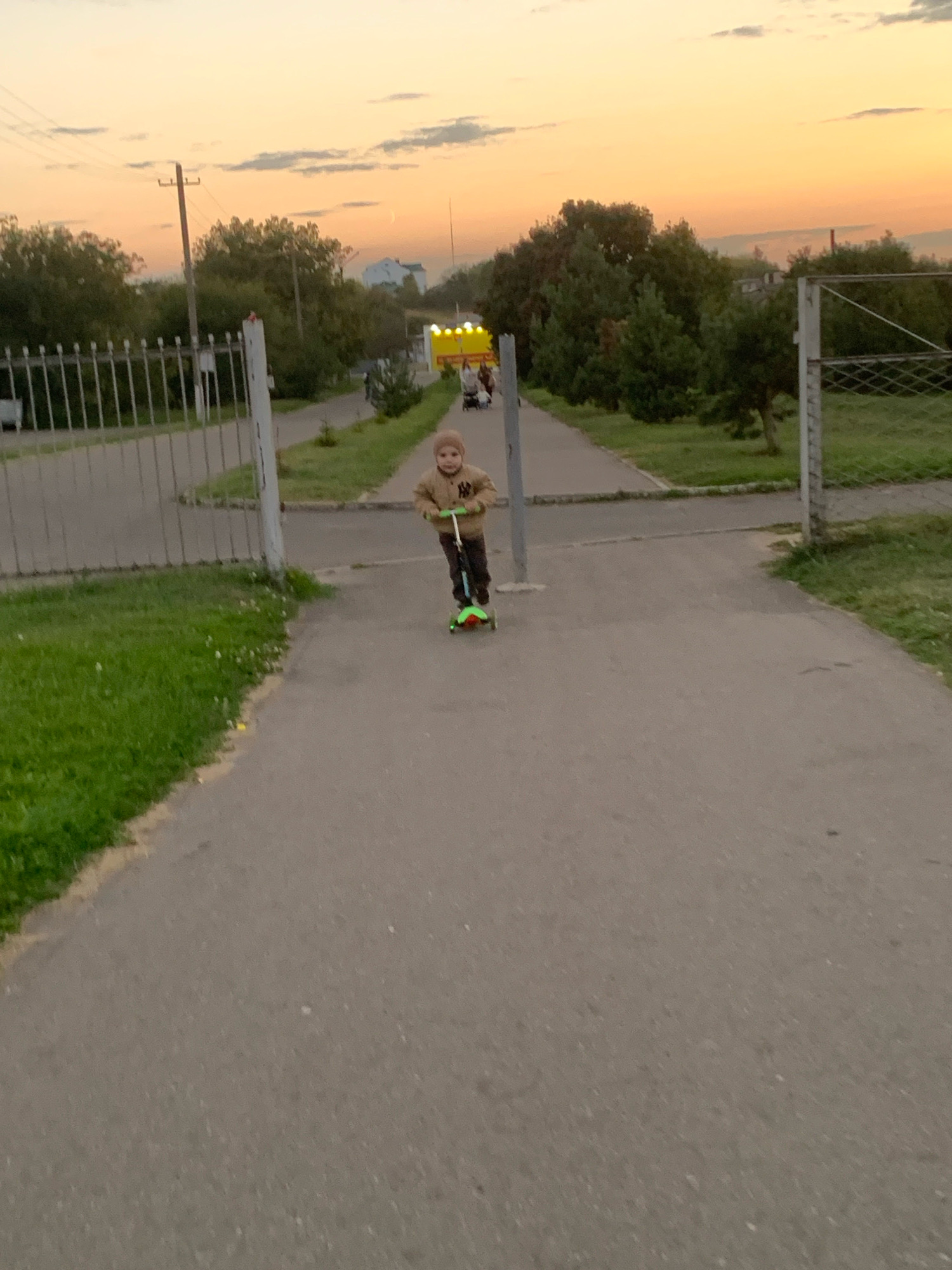 A young child wearing a light brown hooded jacket and dark pants rides a green and black scooter down a paved pathway. The child is facing the camera. The pathway is bordered by a grassy area on the left and a metal fence and more grass on the right. In the background, trees and a yellow structure with lights are visible. The sky above is a soft orange and yellow, suggesting sunset or sunrise. The scene appears to be a park or recreational area in Kapyĺ, Belarus.