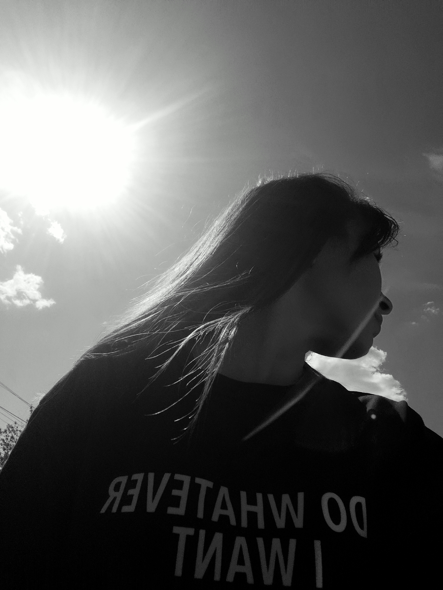 A black and white medium shot captures a person against a bright sky with the sun prominently featured in the upper left. The person, a female, is facing right, her facial features sharply defined against the sun. Her hair is long and straight, catching the light. She is wearing a dark-colored shirt with large, reversed text: "I WANT DO WHATEVER". The background is a blue sky with a few scattered clouds. The location is unknown. There are no visible elements connecting to Фоки, Russia.