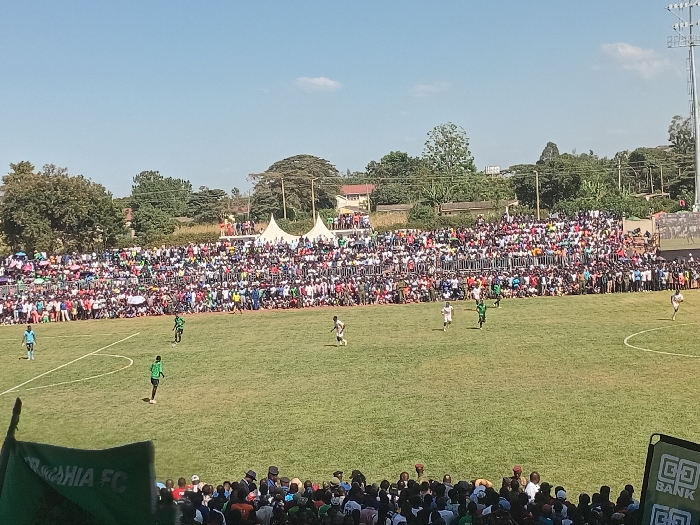 This wide shot captures an outdoor football (soccer) match in progress, viewed from a slightly elevated position within a stadium. The main activity is a game, with several players visible on the lush green grass pitch, distinguished by their green and white kits. A massive crowd, likely thousands, fills the tiered stands that encircle the field, packed tightly together and extending into the distance, suggesting high spectator engagement.
Beyond the densely populated stands, white tents, scattered buildings, and numerous trees are visible, framed by a bright, clear blue sky with only a few wispy clouds. A tall floodlight tower can also be seen on the right side above the stands. The sunny conditions clearly indicate daytime. In the immediate foreground, additional spectators are visible, along with parts of a green banner or flag on the left, displaying the partial text "...AHIA FC." On the right, a black and green sign with the logo and text "GO BANK" is prominently displayed. The scene depicts an energetic sporting event in Siaya, Kenya.