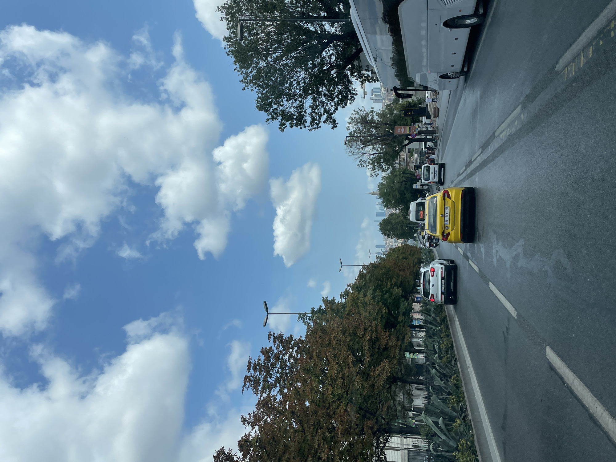 The image depicts a street scene in Istanbul, Turkey, with a focus on the road and vehicles. The sky is partly cloudy, with fluffy white clouds scattered against a blue backdrop. On the right side of the road, a large white bus is traveling in the same direction as the viewer. Several cars are visible on the road, including a yellow taxi in the foreground and a white SUV further ahead. Trees line the left side of the road, some with autumn foliage, and tall lampposts are spaced along the median. In the distance, city buildings and skyscrapers can be seen, indicating an urban environment. The road surface appears wet, suggesting recent rain. There are a few pedestrians visible on the sidewalk in the distance.