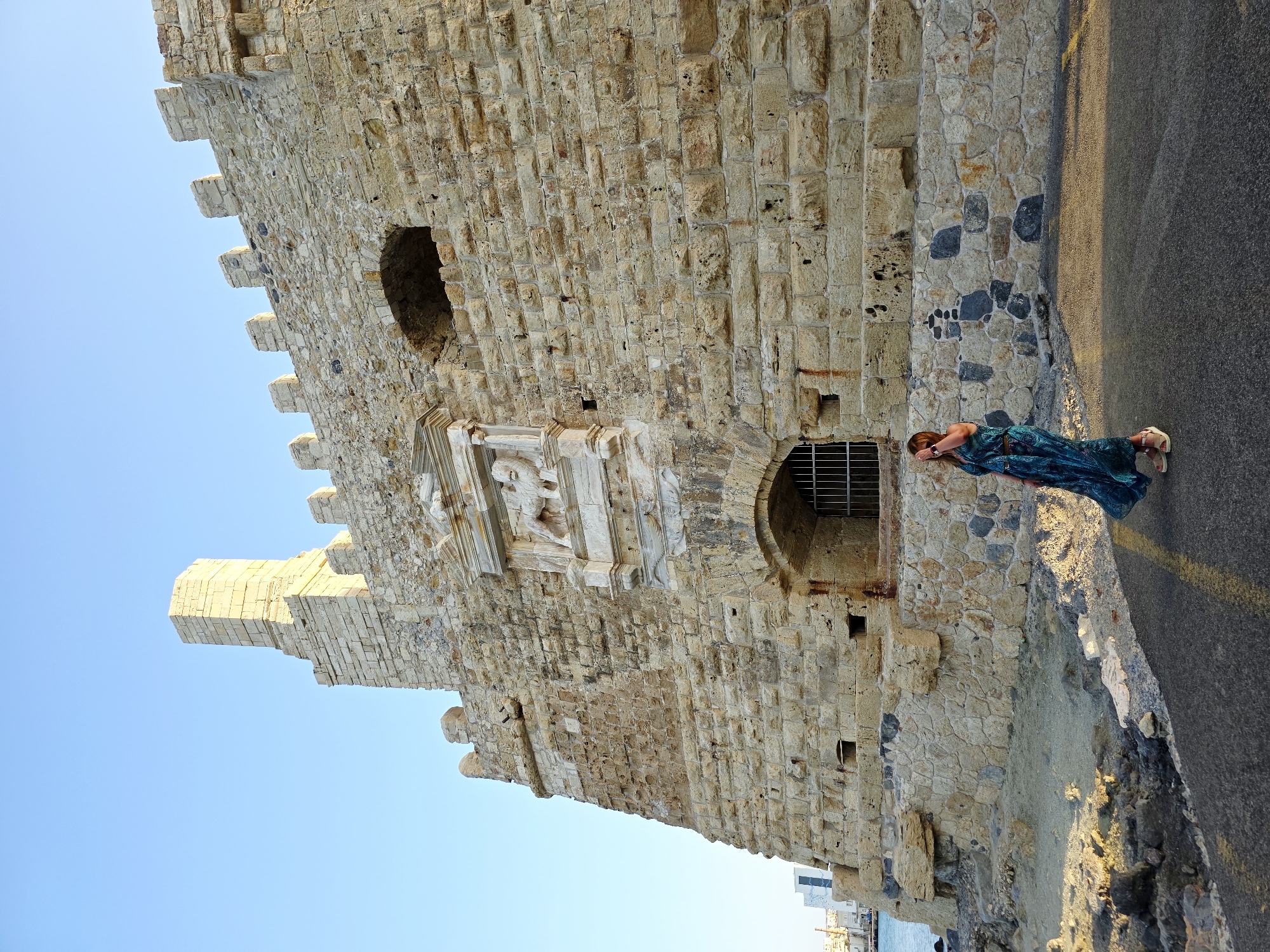 A woman in a long, patterned blue-green dress stands on a paved walkway in front of a large, ancient stone fortress. The fortress is constructed of large, rough-hewn stone blocks and has crenellated battlements along the top. An arched entryway with metal bars is visible on the lower section of the wall, and above it, there is a decorative relief carving depicting a winged lion. The woman is facing left, with her hand touching her hair, and her dress billows out around her. The sun is casting long shadows, indicating it is late afternoon or early evening. The background shows a clear blue sky and a body of water with some distant buildings. This is likely the Koules Fortress (also known as the Rocca a Mare), a Venetian fortress guarding the old port of Heraklion, Crete, Greece.