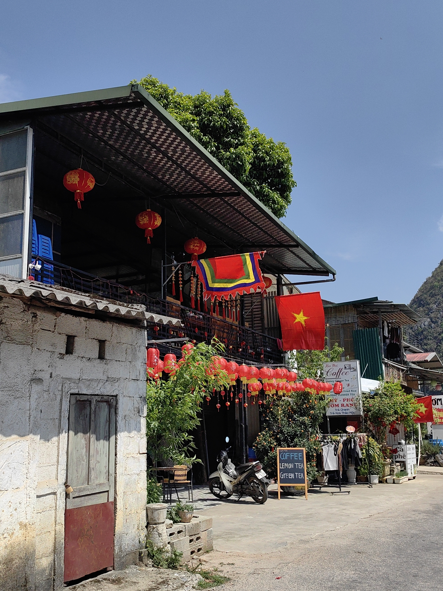 The image shows a street scene in Sà Phìn Commune, Vietnam, on a bright, sunny day. The architecture is a blend of rustic and commercial styles. A two-story building dominates the left side of the frame, adorned with red lanterns strung across the front and hanging from the upper level. Vietnamese flags, including one with a yellow star, are prominently displayed.
A small whitewashed structure with a weathered door stands to the left of the main building. In front of the building, a parked scooter, potted plants, and outdoor seating indicate a café or small business. A chalkboard sign advertises "Coffee, Lemon Tea, Green Tea." Other signs in Vietnamese can be seen, suggesting the availability of food and other beverages. Clothing hangs on a rack, possibly for sale. The street is paved and appears relatively quiet. Beyond the buildings, hints of lush greenery and possibly mountainous terrain are visible.