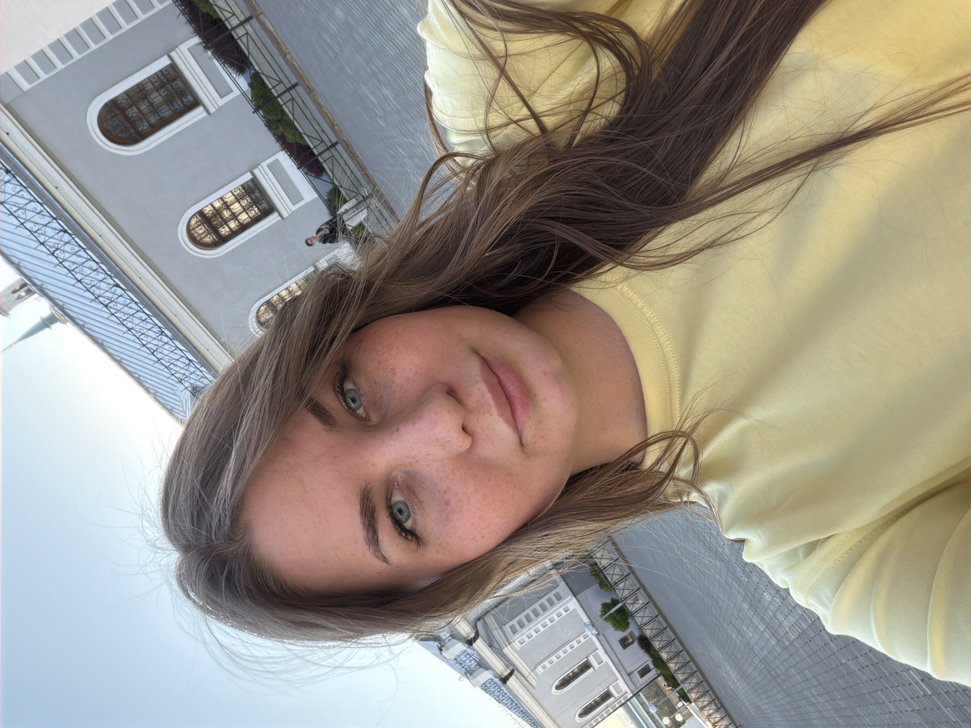 An outdoor, slightly tilted selfie features a young adult female in the foreground, facing the camera. She has long, straight brown hair parted down the middle, blue eyes, and noticeable freckles across her nose and cheeks. She is wearing a plain, light yellow, crew-neck, long-sleeved shirt or sweatshirt.
In the background, a large historical building with a light gray facade, white architectural trim, and multiple arched windows with dark grates or frames is prominent. The building is topped with a distinct metallic blue-gray gabled roof featuring a black decorative railing along its ridge. To the upper left, a slender, light green spire with a golden tip is visible against a clear blue sky. The ground in front of the building is a wide expanse of light gray patterned pavers or cobblestones. A black metal fence with horizontal bars runs along the far edge of the paved area, separating it from landscaped bushes and small trees. Further in the background, to the right, a lone male figure wearing a dark top and light pants stands near the fence.
The architectural style of the building, including the gray facade, white trim, arched windows, distinctive roof, and the visible spire, identifies the location as the Kazan Kremlin in Kazan, Russia. The building in the background appears to be part of the Presidential Palace complex or an adjacent historical structure within the Kremlin, with the spire likely belonging to the Annunciation Cathedral.