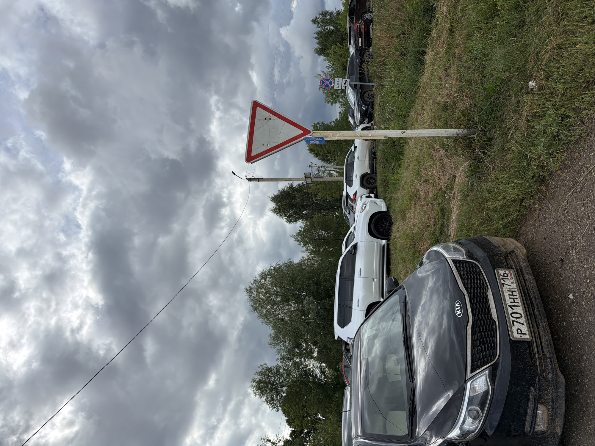 The image captures a parking lot scene under a cloudy sky in Yelabuga, Russia. In the foreground, a black Kia Rio is parked, facing left, with its license plate clearly visible: "P 701 HH 716". Several other vehicles are parked behind it, including a white SUV, a black SUV, and a brown SUV. A white traffic sign, shaped like an inverted triangle with a red border and a white interior, stands on a pole to the right of the road. Behind the vehicles, a line of trees forms a backdrop. A power line is visible stretching across the top of the image.