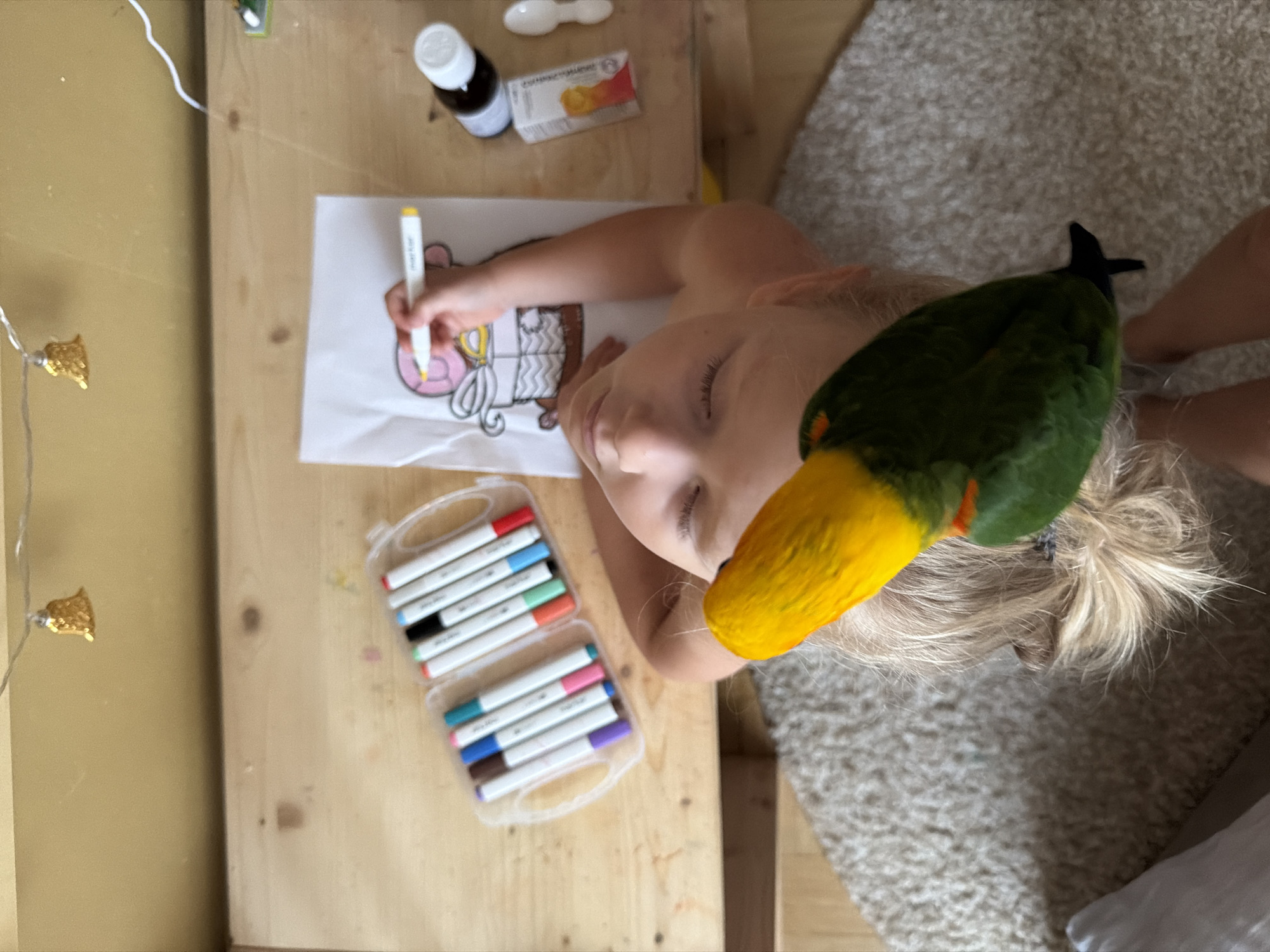 An overhead medium shot shows a young child drawing on a piece of paper with a yellow marker. The child, whose eyes are closed and head is tilted back, has blonde hair that is pulled back in a ponytail. A green and yellow parrot is perched on the child's head, its beak touching the child's forehead. In the background, a set of colorful markers in a plastic case, a small bottle, and a white spoon are on a wooden table. The scene is set indoors, likely a home environment. There is no indication of the city of Khimki, Russia.