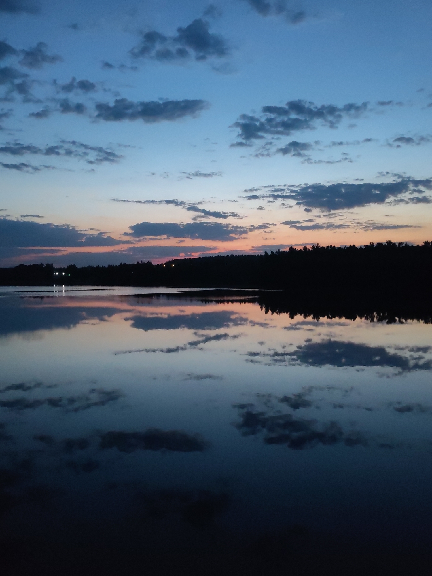 The image captures a twilight scene at a body of water, possibly a lake or a wide river, reflecting the sky. The sky displays a transition from blue at the top to shades of orange and pink near the horizon. Numerous clouds are scattered across the sky, mirrored in the water's surface. A dark treeline is visible along the horizon, with faint lights suggesting a distant settlement. The location is possibly Ermolino, Russia. The image depicts a tranquil, natural scene without the presence of people.
