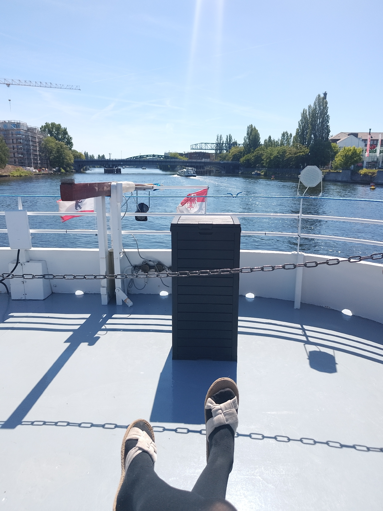 View from a boat traveling on a river. Flags of Berlin and Brandenburg are visible. Person in the foreground wearing black leggings and beige sandals. A bridge is in the background. Crane and buildings under construction on the left. Trees and buildings on the right. The water is blue and reflective. The deck of the boat is grey with a dark grey storage bin.
