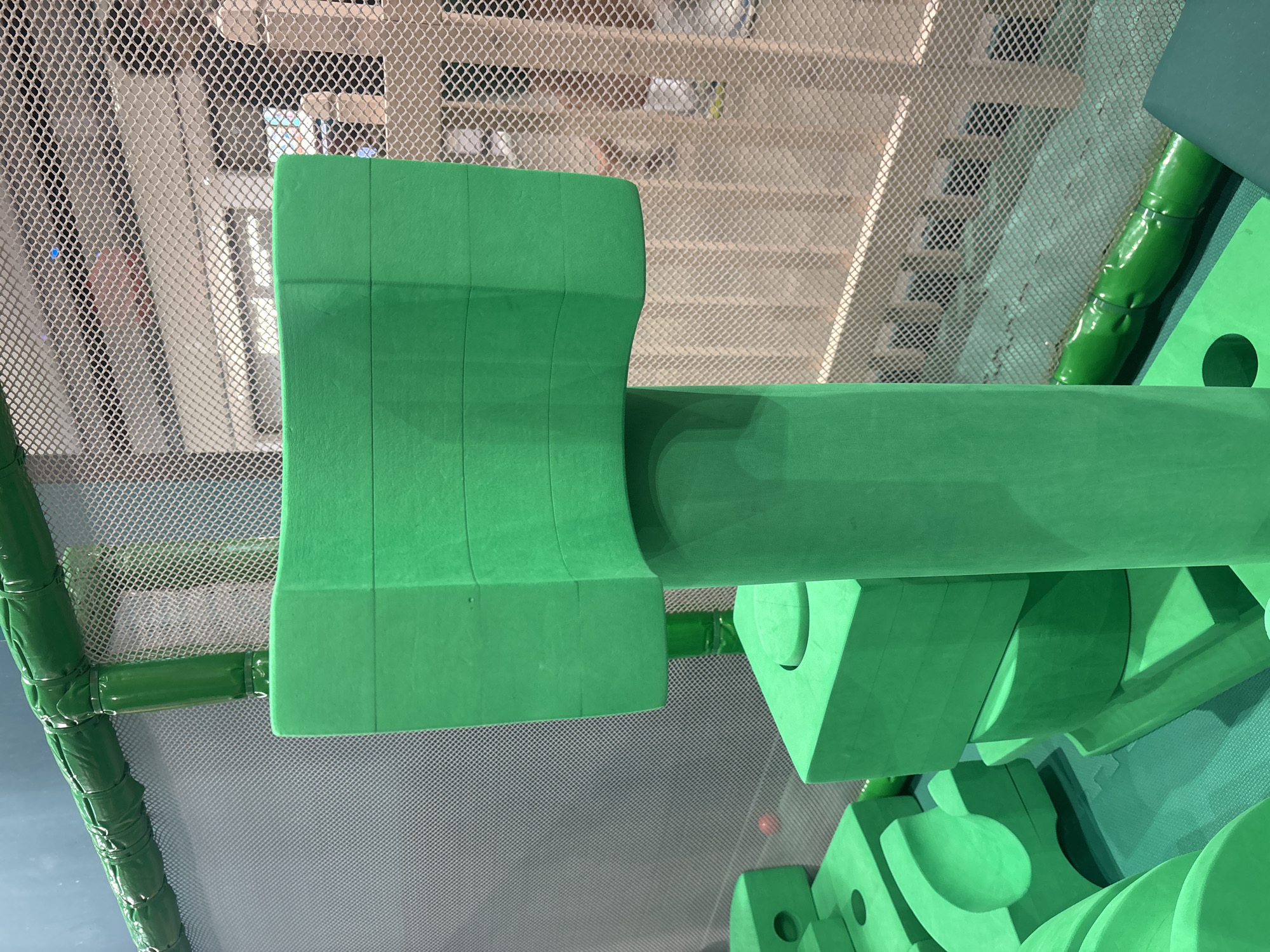 The image depicts a segment of a children's indoor playground, characterized by brightly colored, foam-padded structures. The dominant color is a vibrant green, forming various geometric shapes such as cubes, cylinders, and rounded blocks. These elements appear to be part of a climbing or obstacle course. A mesh netting, likely for safety, forms a backdrop, and beyond that, glimpses of a beige, slatted wall and further structures can be observed. The lighting suggests an indoor setting, possibly a commercial establishment. There are no people or animals visible in the frame. The scene is static and does not show any activity. The image is focused on the play structures, highlighting their textures and forms. There is no discernible information within the image to indicate a specific location in Dubai, United Arab Emirates.