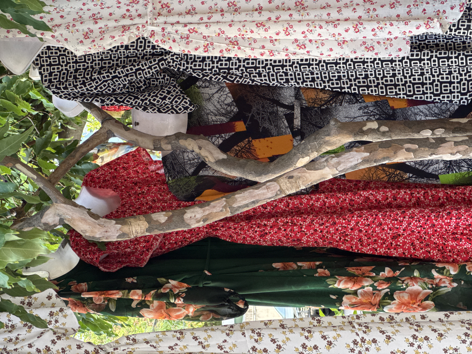 A street market stall in Nha Trang Ward, Vietnam, displays several colorful dresses for sale, hanging from branches of a tree. The dresses feature various floral and geometric patterns. On the left, a white dress with small brown and yellow flowers is visible. Next to it, a dark green dress is adorned with large, vibrant orange and pink flowers. To the right of the green dress, a red dress showcases a dense pattern of tiny white flowers. Further to the right, a black and white dress has a repeating geometric pattern. The last dress on the far right is white with small red and yellow flowers. The dresses are displayed outdoors, with lush green leaves from the tree overhead providing shade. The background suggests an outdoor setting, possibly a market or street. No people are visible in the frame.