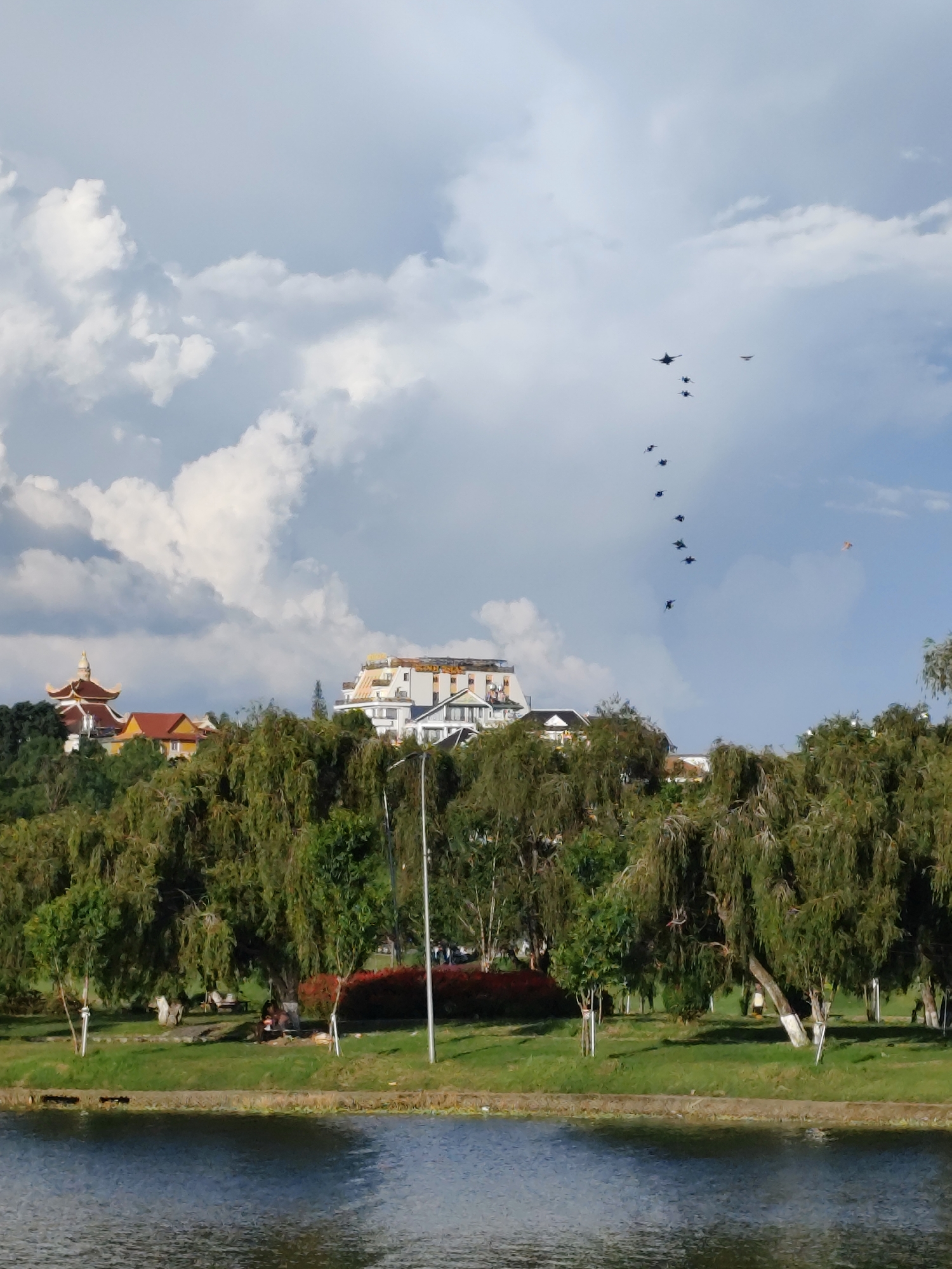 The image depicts a scenic view of Xuan Huong Lake in Da Lat, Vietnam, under a partly cloudy sky. In the background, several buildings are visible on a hillside, including a prominent structure with a golden-tiled roof that resembles a pagoda, and a larger, modern building with white and yellow facades. A flock of at least ten dark-colored aircraft, appearing to be fighter jets, are flying in a V-formation across the sky. Closer to the viewer, a line of lush green trees with drooping branches forms a natural boundary along the lakeshore. The water of Xuan Huong Lake occupies the foreground, reflecting the sky and surrounding trees. On the grassy bank, a few people are visible sitting or walking, and a lamppost stands near the treeline. The overall impression is a blend of natural beauty, urban architecture, and an aerial display.