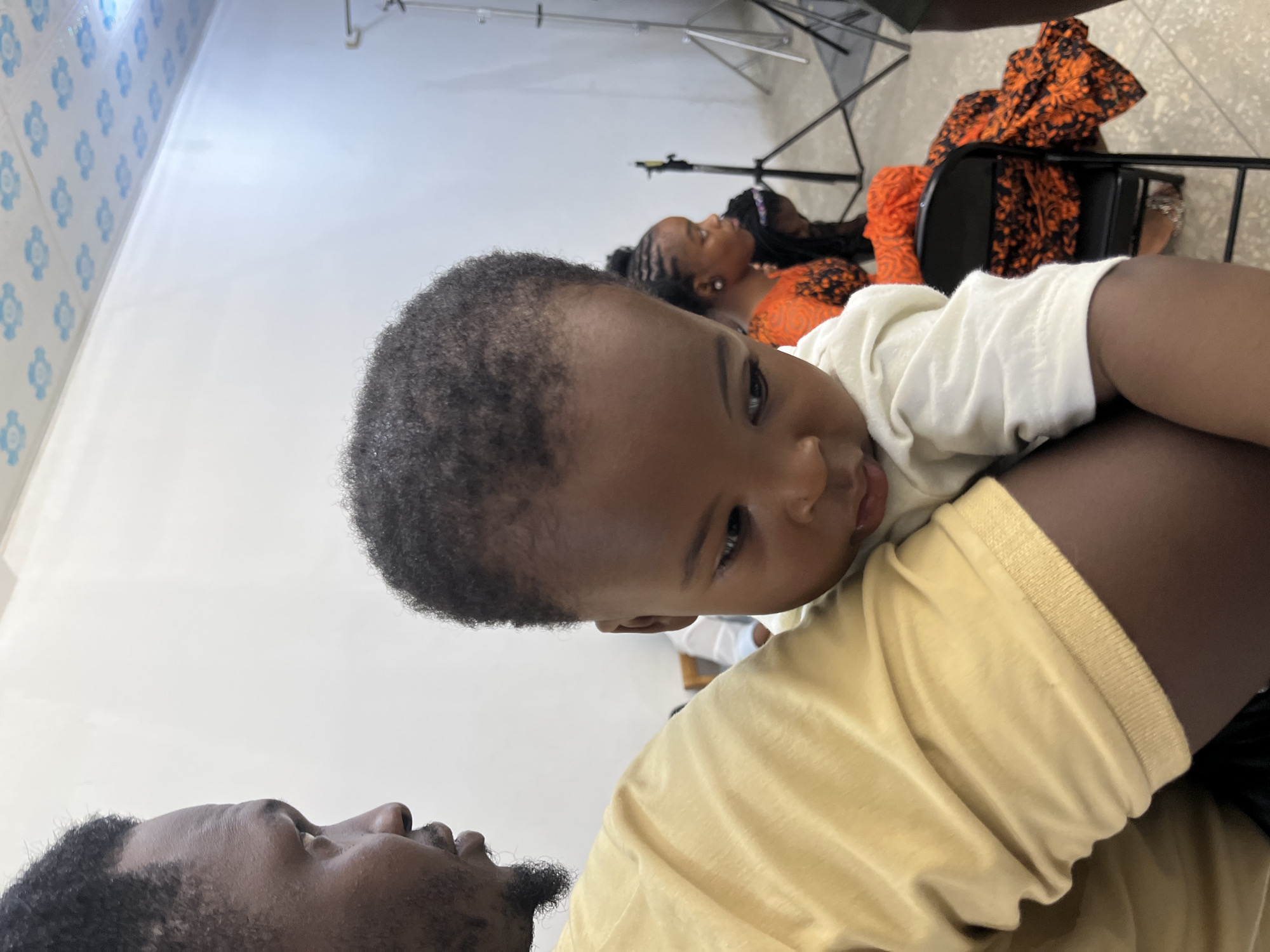 The image shows a close-up of a Black man holding a young Black child. The man is wearing a light yellow shirt and the child is wearing a light-colored onesie. The man is looking to the left of the frame, while the child is looking down. In the background, two other Black individuals are visible. One is a woman wearing an orange and black patterned garment, and another person is partially visible behind her. The environment appears to be indoors, possibly a studio or event space, with a white wall and a patterned ceiling visible. Lighting equipment stands are also present in the background, suggesting a staged setting. There is no information in the image to indicate the location is Gura Top, Nigeria.