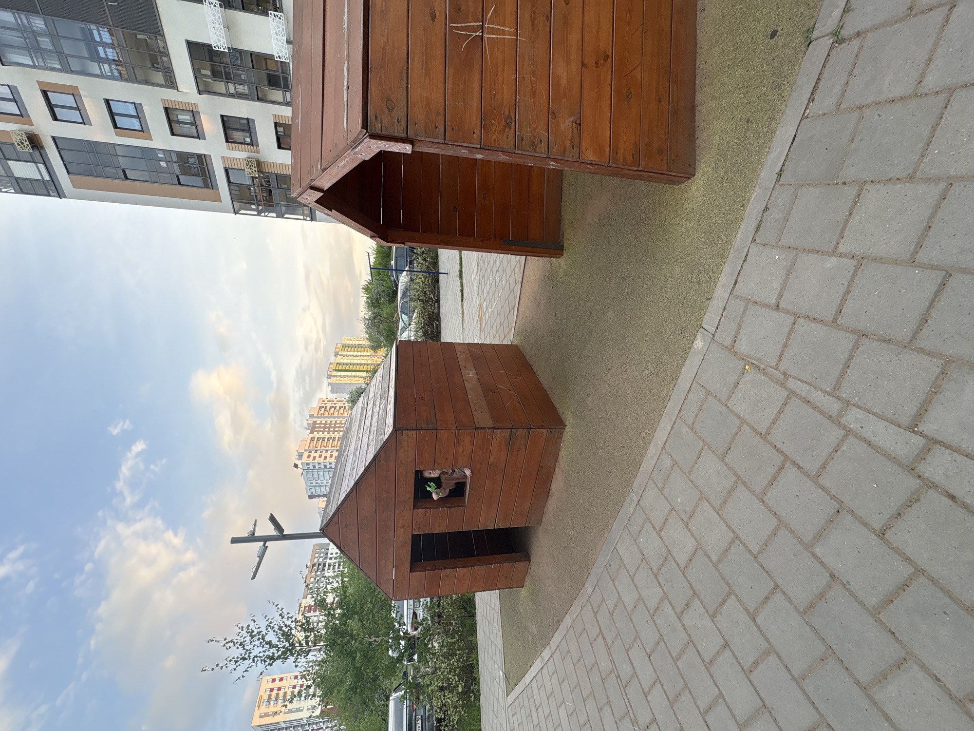 The image depicts a small wooden playhouse on a playground in an urban area, likely in Izhevsk, Russia, judging by the architectural style of the surrounding buildings. The playhouse consists of two separate structures, one a simple enclosure and the other a small house with a window and door. The playground is situated on a paved area with gray concrete pavers, and there is artificial turf surrounding the playhouse. The background features modern residential buildings, typical of contemporary urban development in Russia. A child is visible inside the playhouse, looking out the window. A streetlight post is also visible in the background.