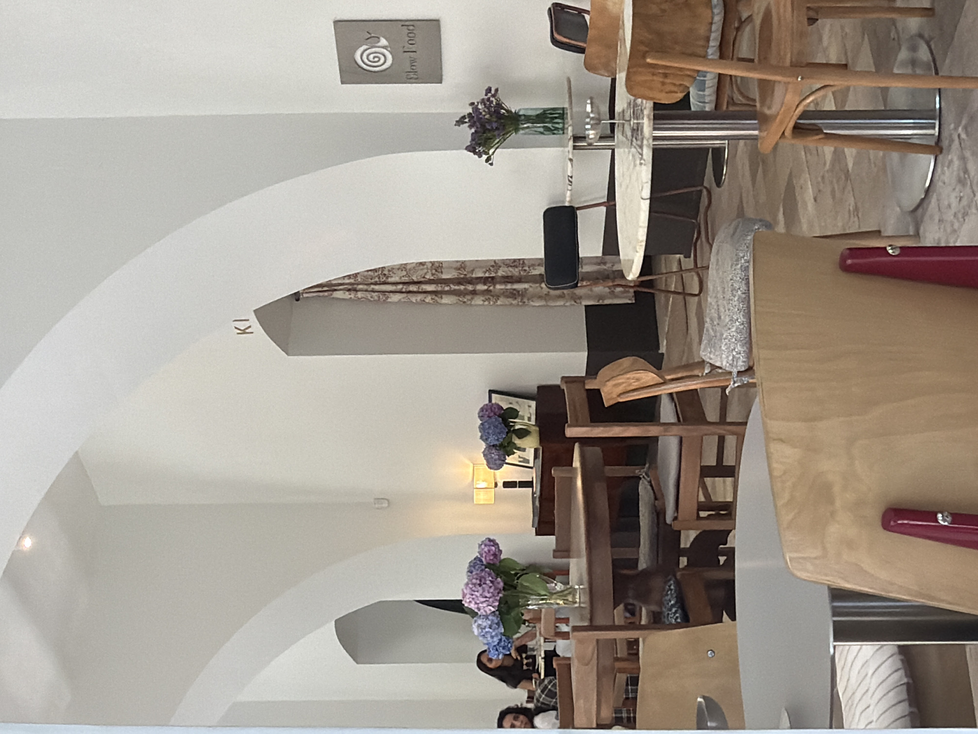 This image depicts the interior of a cafe with a minimalist design, featuring white arched ceilings and light-colored walls. Several tables and chairs are arranged throughout the space, indicating it is a seating area. On one table, there is a vase with purple and blue hydrangeas, and on another, a vase with purple flowers. A lamp is visible on a dark wood cabinet, next to a framed picture. A sign with a snail logo and the words "Slow Food" is mounted on the wall. The cafe appears to be located in Baku, Azerbaijan, as suggested by the presence of the "Slow Food" organization which is known to have chapters in Azerbaijan. The overall ambiance is calm and inviting, suitable for dining or socializing.