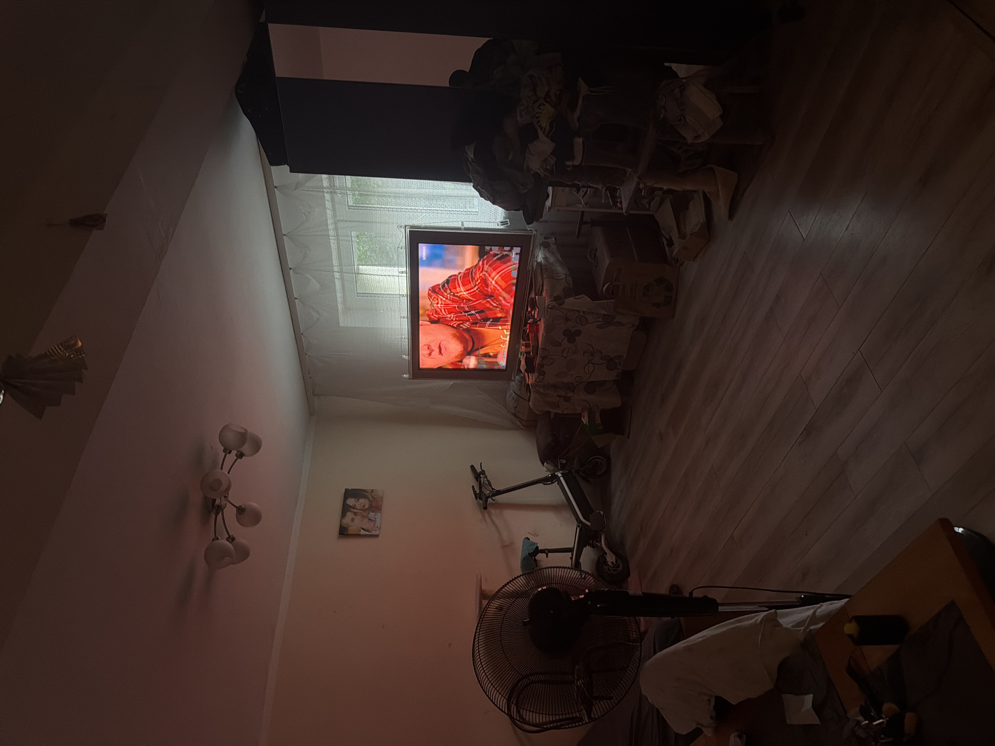 The image depicts a dimly lit room, likely a living area, with wooden flooring. A large, dark-colored fan is positioned on the left, partially obscured by a white cloth draped over what appears to be a table or surface. A black electric scooter is parked in the middle of the room. A television screen displays a person wearing a red and black plaid shirt. On the wall above the scooter, a framed photograph of two people is visible. The room contains a window covered by sheer white curtains. To the right of the television, a dark wardrobe or cabinet stands, with items of clothing or fabric piled on top. There is a shelf unit near the wardrobe, also with items on it. The overall impression is one of a lived-in space, with personal belongings scattered throughout. The lighting suggests it is either evening or the room has limited natural light. There is no specific information within the image to suggest the location is Szczecin, Poland.