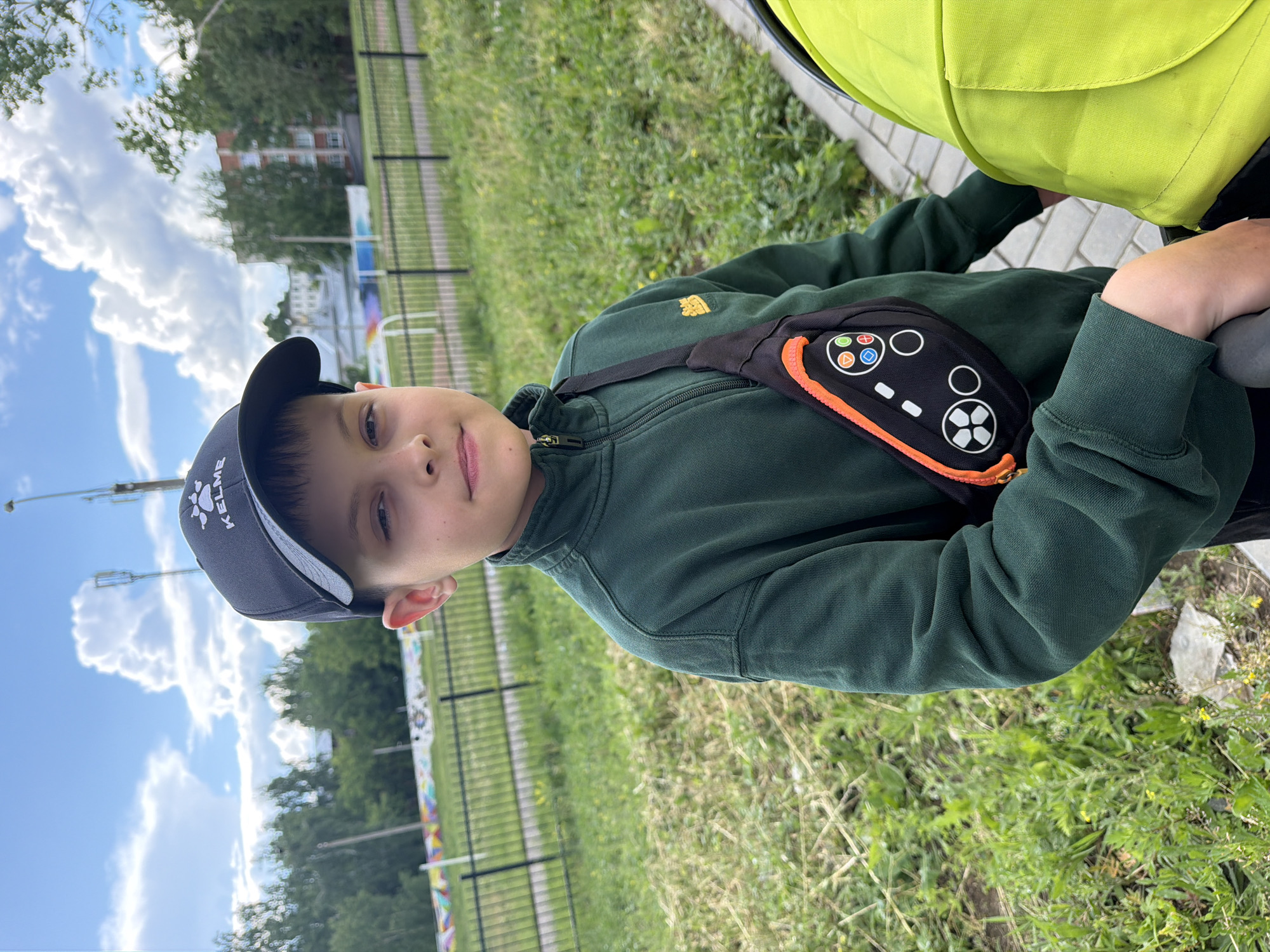 A young boy, appearing to be of elementary school age, is captured looking directly at the camera with a calm expression. He is wearing a dark grey baseball cap with the white "KELME" logo on the front and a dark green zip-up jacket. A black fanny pack, designed to resemble a video game controller with a prominent orange zipper, multi-colored buttons (including red, blue, green, yellow, and white symbols), and a white directional pad, is worn across his chest. To his right, a section of a bright lime-green fabric item, likely part of a baby stroller or pram, is visible.
The scene is set outdoors on a bright, partially cloudy day. The background features a lush green grassy area, a black metal fence, and numerous trees. In the distance, several light-colored buildings can be seen. The sky is a clear blue with large, white cumulus clouds. The image conveys a casual, outdoor moment.