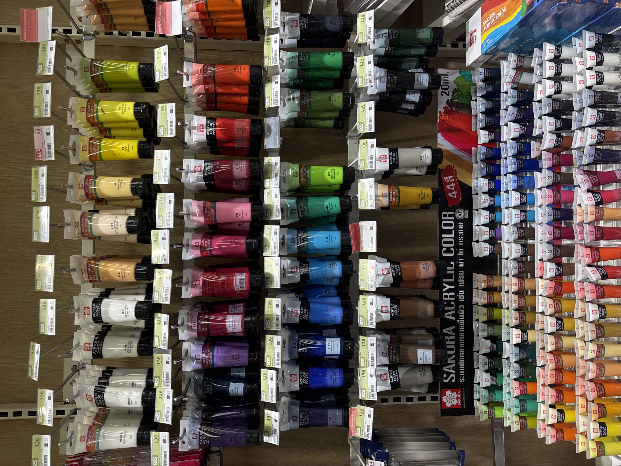 The image shows a well-lit, orderly display of Sakura Acrylic Color paint tubes in a retail setting. The scene is likely inside a shop, possibly an art supply store, given the merchandise. The tubes, neatly arranged on metal shelving units with individual hooks or pegs, form vertical rows of varied colors. Each tube is individually packaged in clear plastic with a price tag and barcode. The "B2S" branding appears on price tags, possibly indicating the retail chain. A promotional sign for "Sakura Acrylic Color" in Thai and English, featuring the brand's cherry blossom logo, is situated centrally. The shop's location is suggested to be in Choeng Thale, Thailand.