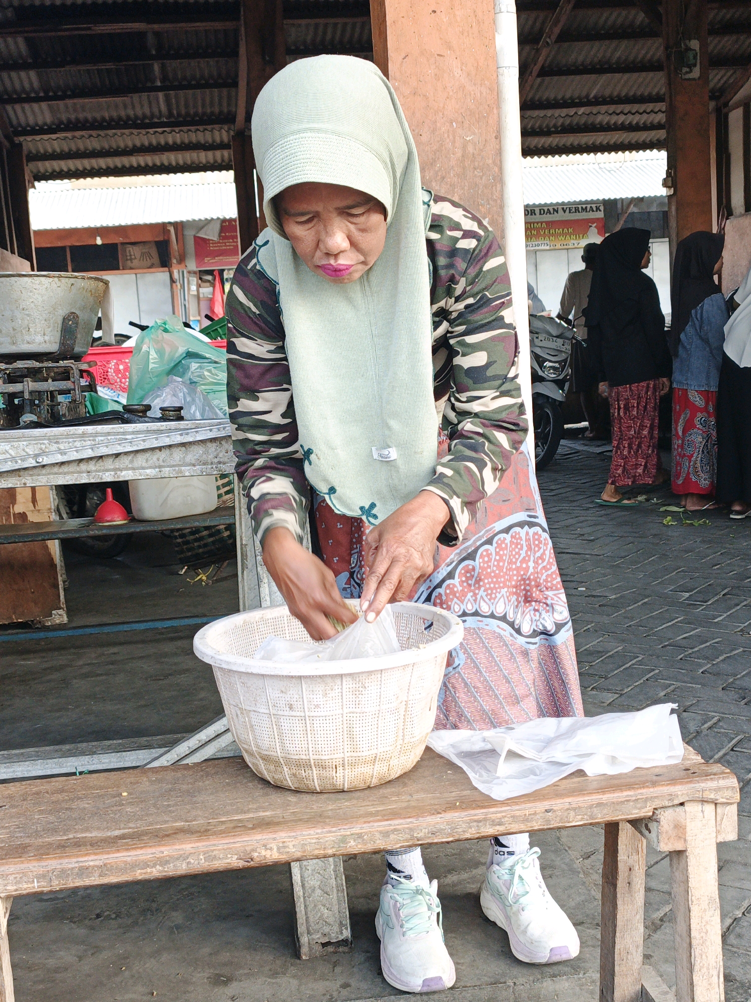 In Kludan, Indonesia, a woman wearing a light green hijab and a camouflage patterned long-sleeved shirt is seen from the waist down, in front of a stall. She is looking down at a white woven basket in front of her. She has on a patterned skirt and white sneakers with light blue accents. A clear plastic bag is on the wooden bench next to the basket, and her hands are reaching into the basket. Behind her, a motorcycle is visible, as are several other people in headscarves, some wearing long patterned skirts. The ground is paved with dark gray interlocking bricks. The background shows the corrugated metal roof of a structure, supported by wooden beams. The stall appears to be set up in an outdoor marketplace.