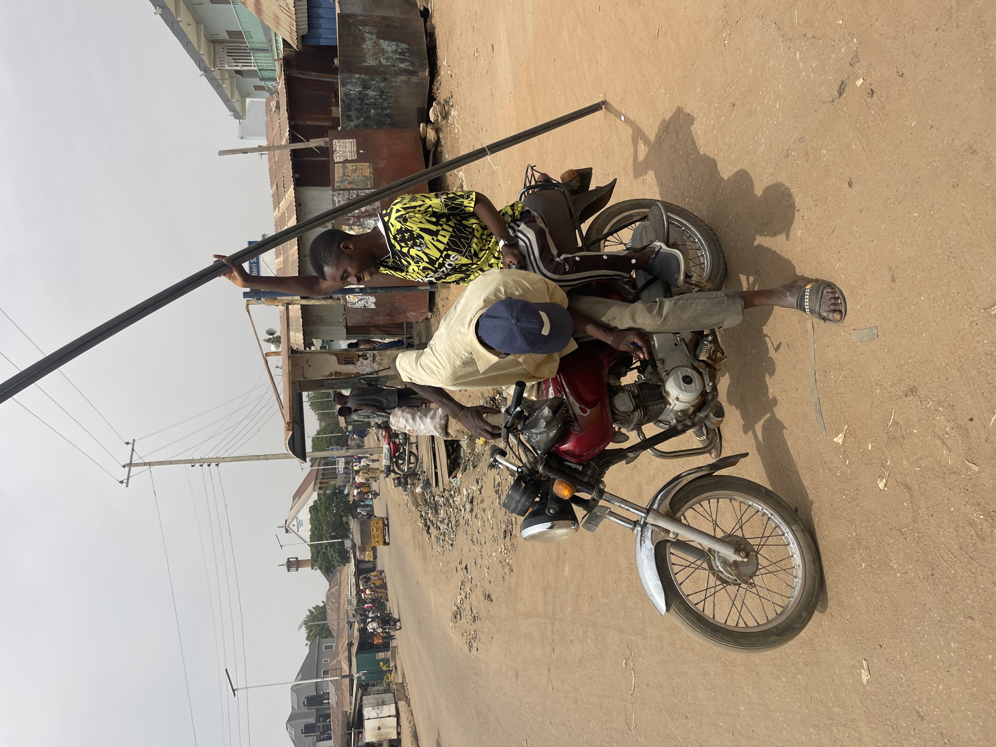 In Chikaji, Nigeria, two men are working on a red motorcycle parked on a dusty street. One man, wearing a light yellow shirt and a blue cap, is bending over the engine. The other man, wearing a yellow and black patterned shirt and dark pants with stripes, is holding a long, dark metal bar above the motorcycle. The street is lined with buildings, including a dilapidated metal structure and a building with a corrugated roof. Power lines are visible in the background. The street is filled with other motorcycles and people, suggesting a busy urban environment. The overall impression is one of everyday life and improvised repair in a bustling city.