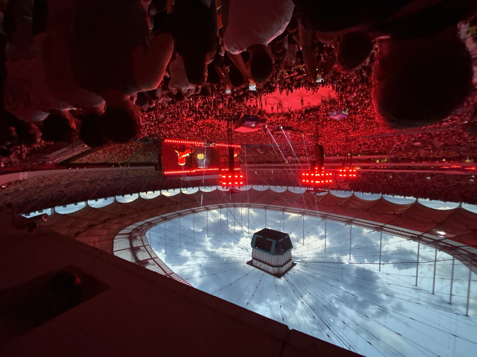 A crowded stadium is illuminated in red light, with a concert stage and screens visible. The view is from a high vantage point looking down at the crowd and stage. Above the stadium, a retractable roof is partially open, revealing a cloudy sky. The location is likely a large sports arena or concert venue in Unknown, Unknown.