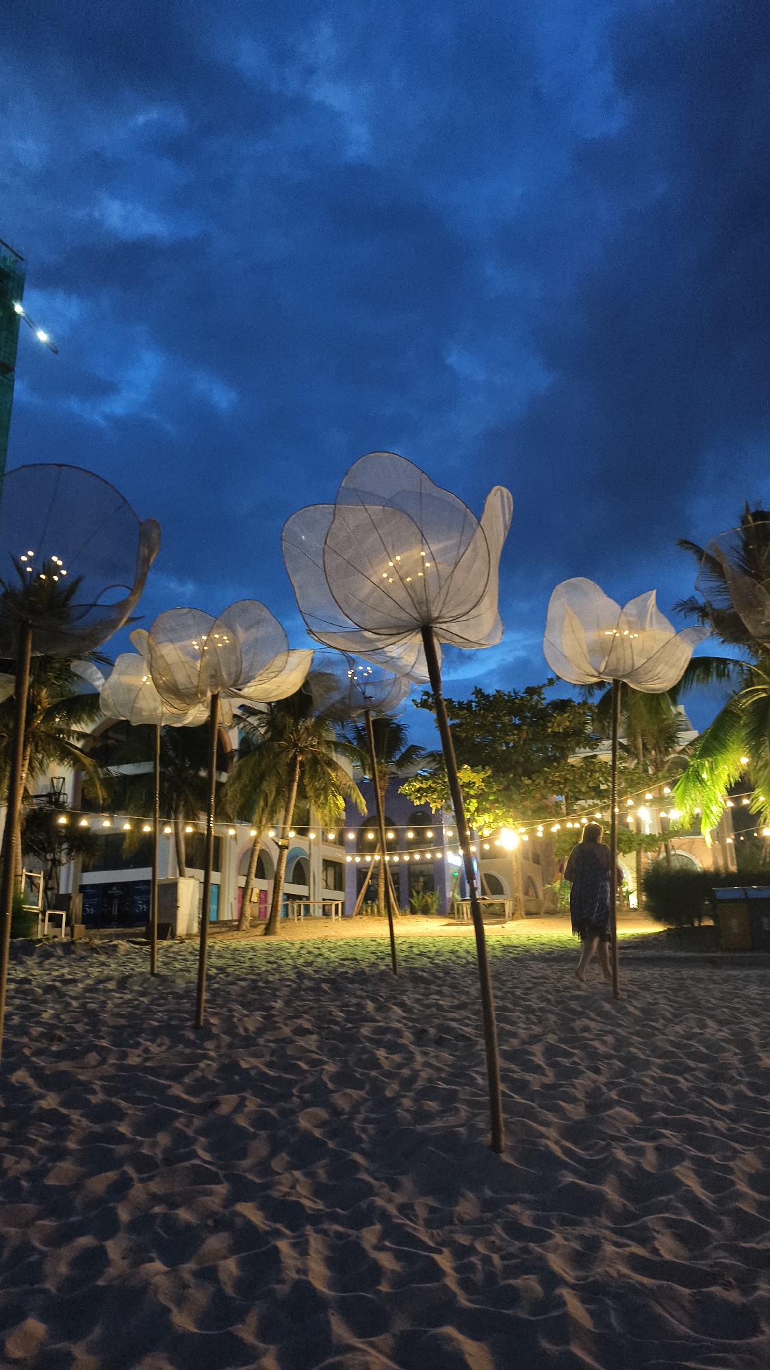 A vertical, low-angle shot captures a beach at dusk in Nha Trang, Vietnam. The sand in the foreground is textured with shadows cast by several large, white, flower-shaped lights mounted on wooden poles. These lights, made of a translucent material and illuminated from within, are arranged across the scene. In the mid-ground, a person wearing a dark, patterned dress walks away from the camera towards the right. Behind them, palm trees and a building with arched doorways are visible, decorated with a string of warm, yellow lights. The sky above is a deep, cloudy blue.