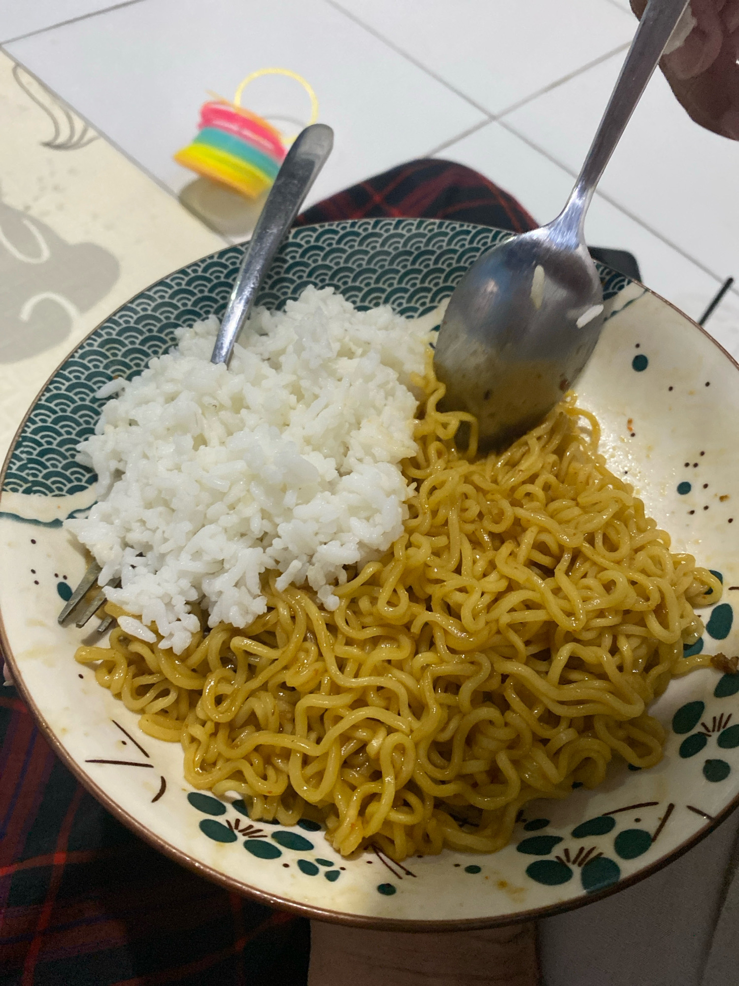 The image shows a close-up of a bowl of food, likely instant noodles with rice, in what appears to be a domestic setting in Kedung Sukodani, Indonesia. A fork is partially visible on the left side of the bowl, and a spoon is held by a hand in the upper right corner, with rice on the spoon. The bowl itself has a decorative pattern of dark green waves on the rim and scattered small dark green shapes and dark brown lines. A colorful, coiled object, possibly a toy or hair accessory, is visible in the blurred background on the left. The food is arranged with white rice piled on the left and golden-brown noodles on the right. A checkered fabric, likely clothing or a tablecloth, is visible beneath the bowl.