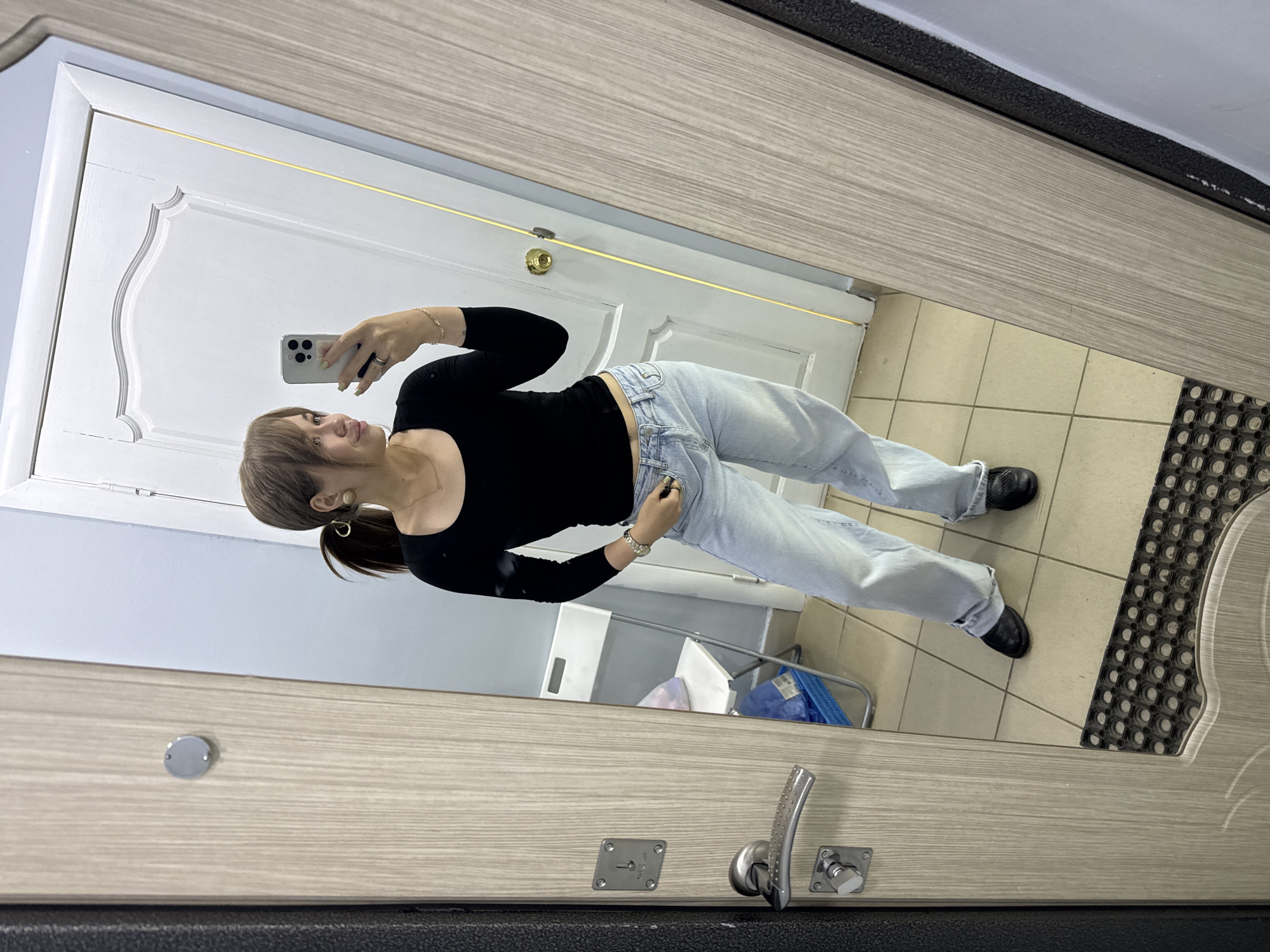 A young woman with brown hair in a ponytail, wearing a black long-sleeved shirt and light-wash jeans, is taking a mirror selfie in what appears to be a hallway. She is standing in front of a light-colored door with decorative molding. To her left is a wooden door frame with a metal door handle and lock plate. The floor is tiled in a neutral color, and a black rubber mat is visible in the lower portion of the frame. In the reflection, a white folding chair and a blue basket are partially visible behind the woman. The overall setting suggests an indoor residential or utility space. The image was captured in Chaikovsky, Russia.