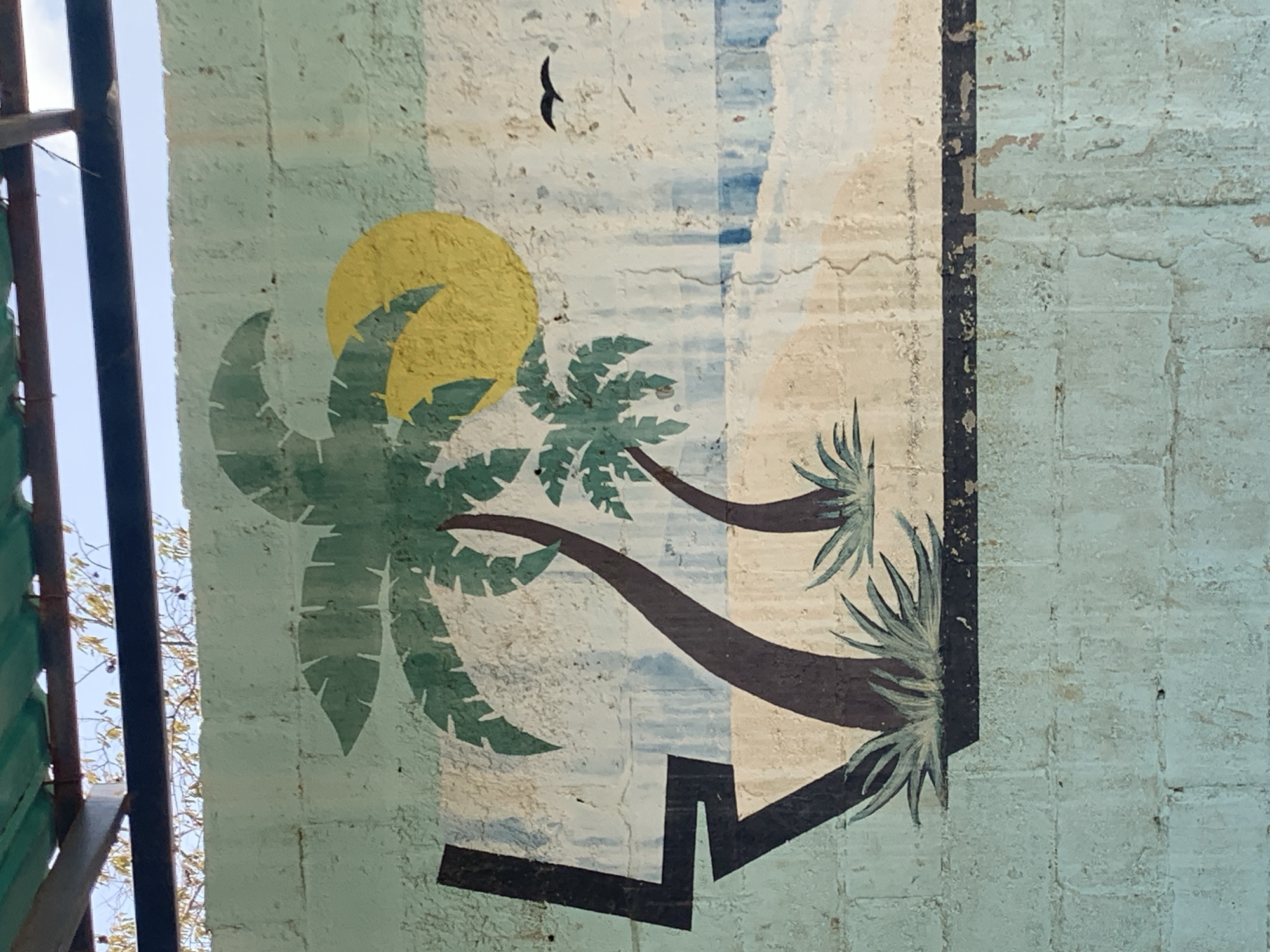A close-up, slightly tilted, eye-level shot shows a painted mural on a weathered, light-blue, textured concrete wall, partially framed on the left by a dark metal structure. The mural depicts two stylized palm trees against a background of light blue, beige, and white sections, representing possibly the sky, clouds, and sand. A yellow circle represents the sun, partially obscured by the leaves of one tree. A black, silhouetted bird is visible near the top of the mural. The base of the painted scene is defined by a dark, angular line, possibly representing the ground. The mural appears to be aged and slightly damaged, with some paint peeling and flaking. The location, Santa Ana, Venezuela, is not apparent from the image.