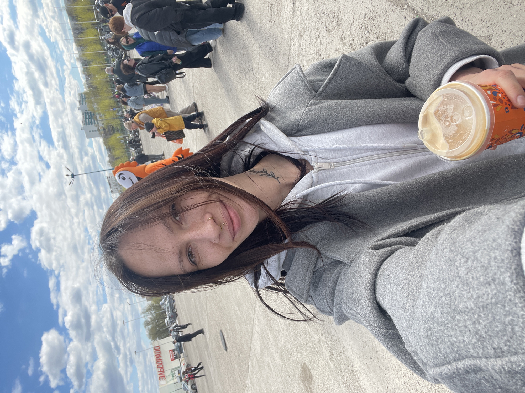A selfie taken at a downward angle captures a young woman in Naberezhnye Chelny, Russia. She wears a gray hooded sweatshirt under a gray coat and has a nose ring and neck tattoo. She holds a cup of coffee. The background shows a cloudy sky, a crowd of people, and a building with the Cyrillic text "ЭЛЬДОРАДО" visible on it.
